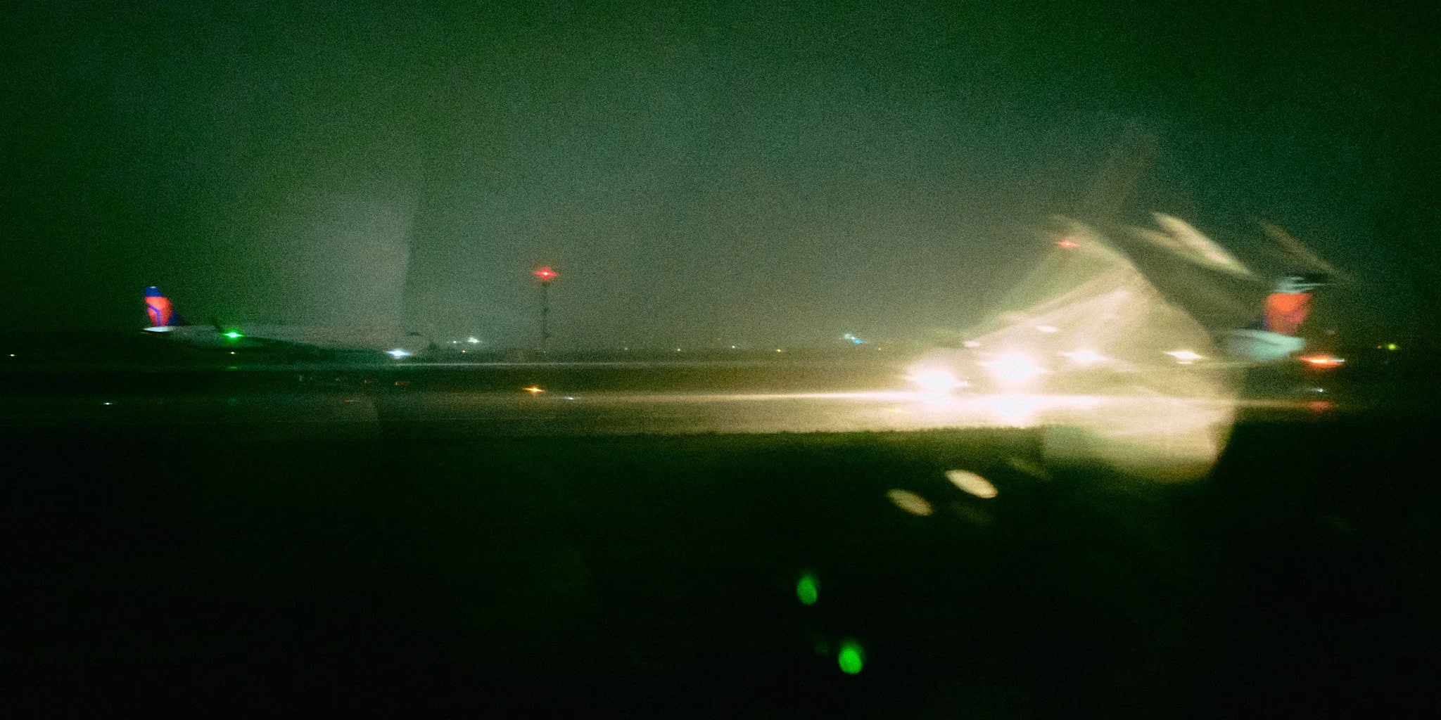 Nighttime shot of an airplane on the runway, with bright lights reflecting off the wet surface and some red and green airport lights visible in the background.