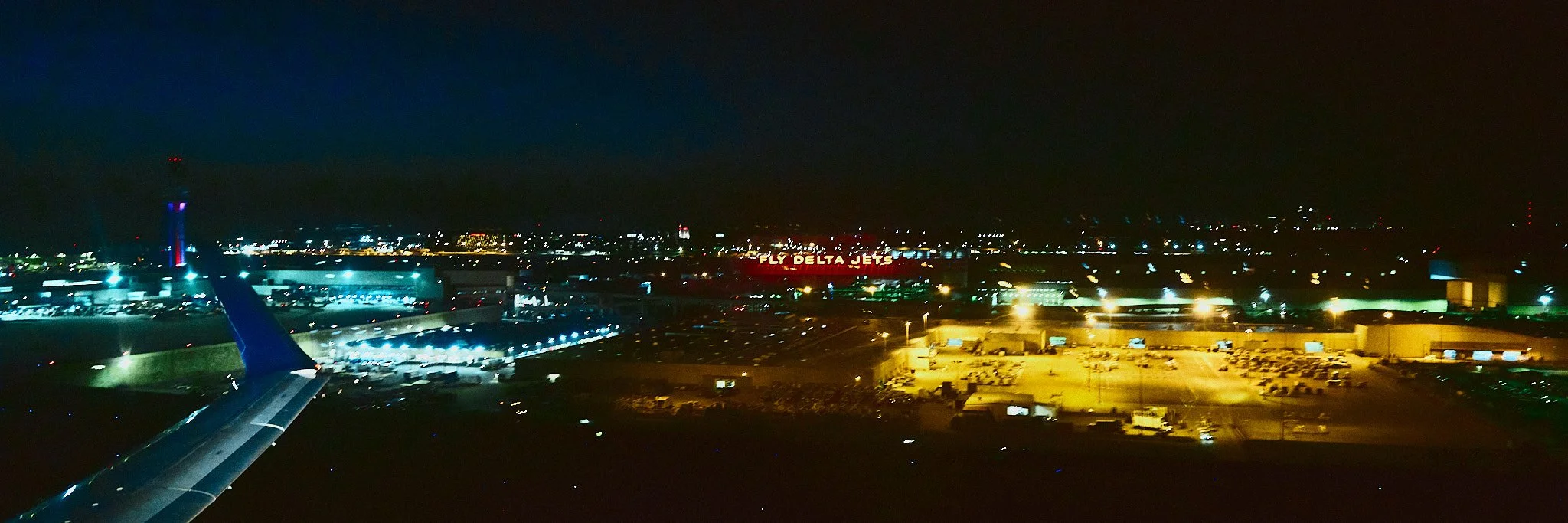 Nighttime aerial view of an airport with illuminated runways, aircraft, terminals, and surrounding city lights. Visible signage includes 'Fly Delta Jets'.