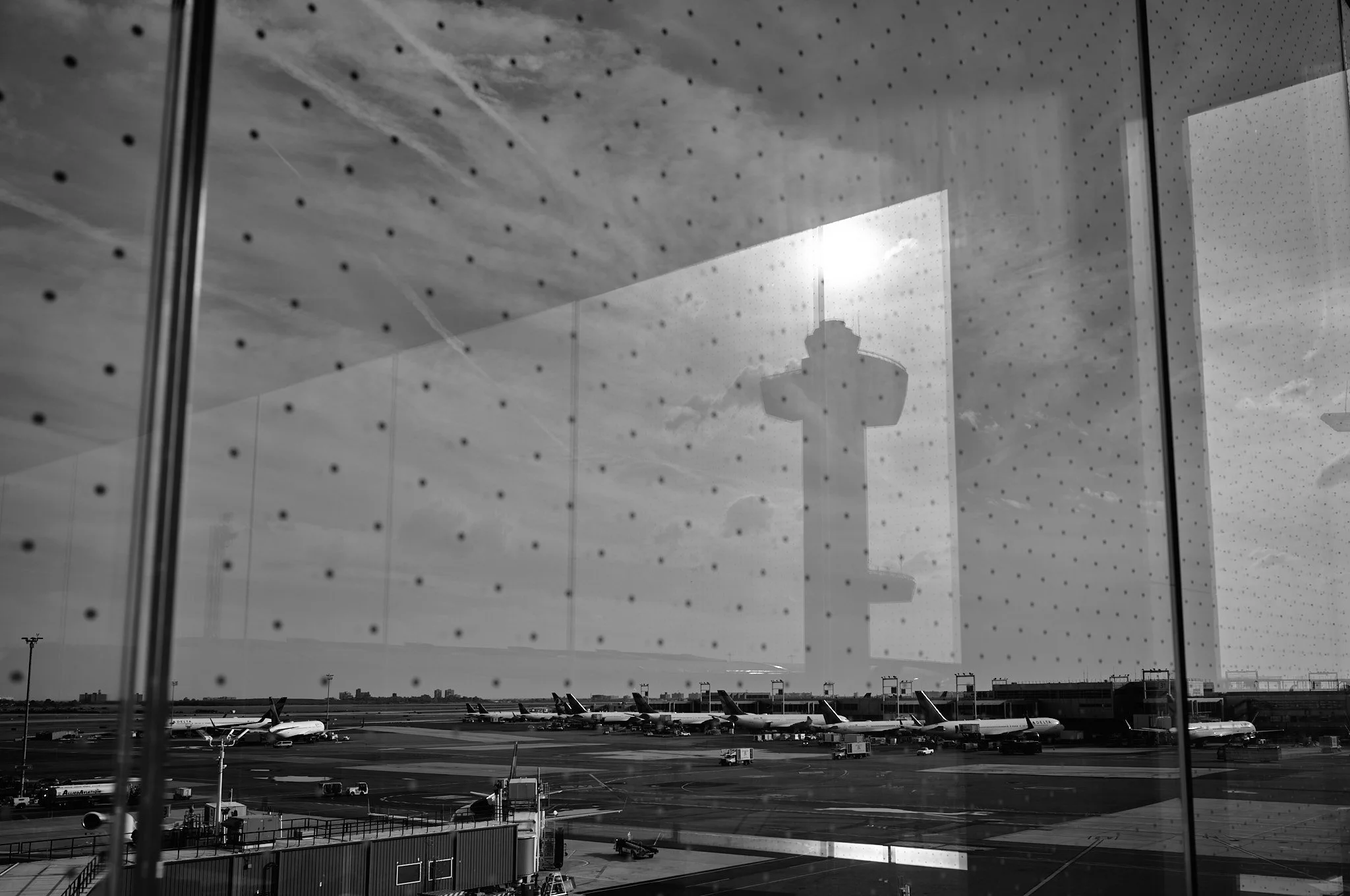 View of an airport tarmac with airplanes, seen through a large window with a dotted pattern. The reflection of an airport control tower is visible in the window, with the sun shining behind it. The scene is in black and white 
 by Matt Ellis