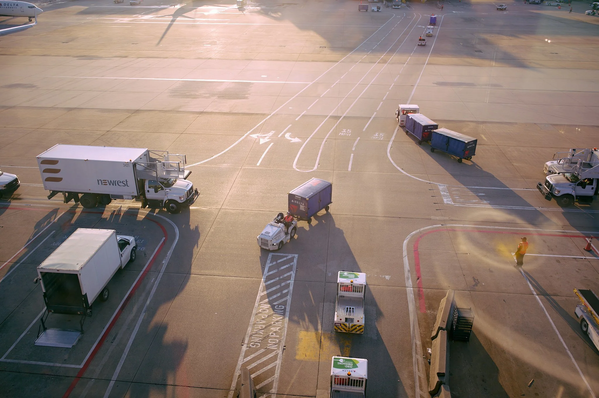 View of an airport tarmac with various aircraft ground support vehicles and equipment, including baggage carts, tug vehicles, and service trucks, with a Delta aircraft partially visible in the upper left corner  by Matt Ellis