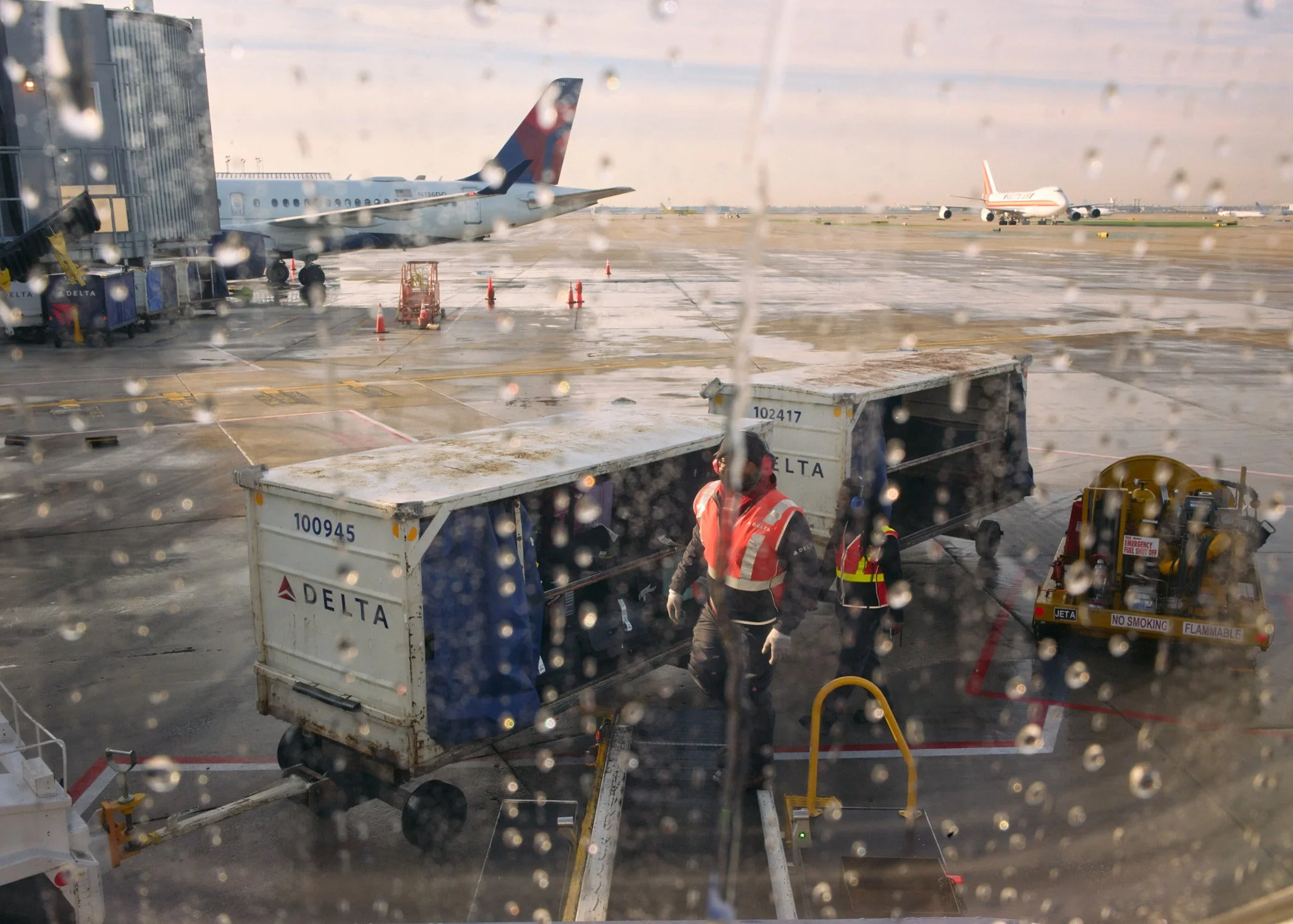 View of an airport tarmac seen through a rain-speckled window, with two Delta aircrafts parked at gates, ground crew working near baggage carts, and a rainy sky in the background  by Matt Ellis