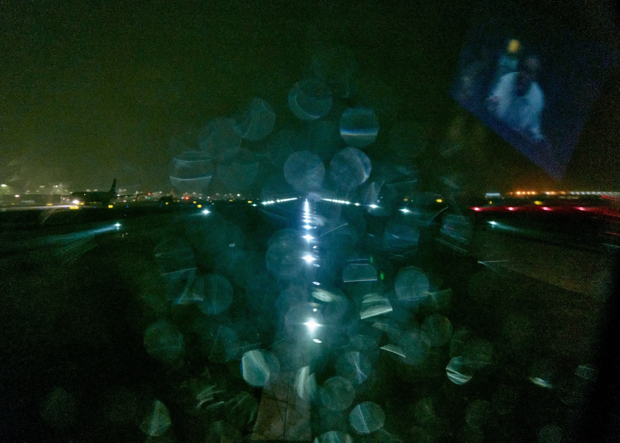 Nighttime runway scene viewed from an airplane window with rain droplets on the window, obscuring the view of the runway lights and a plane parked on the tarmac  by Matt Ellis