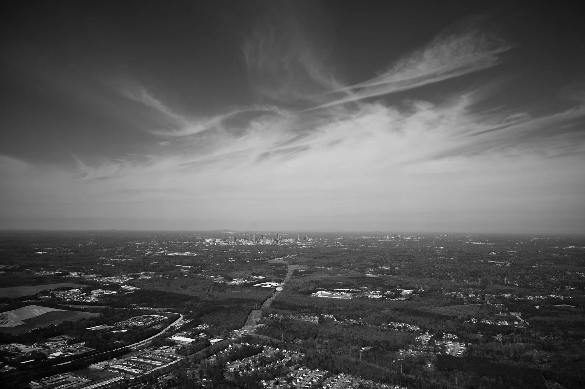 Black and white aerial photograph of a cityscape with a distant skyline, surrounded by suburban areas and open land, under a sky with wispy clouds  by Matt Ellis