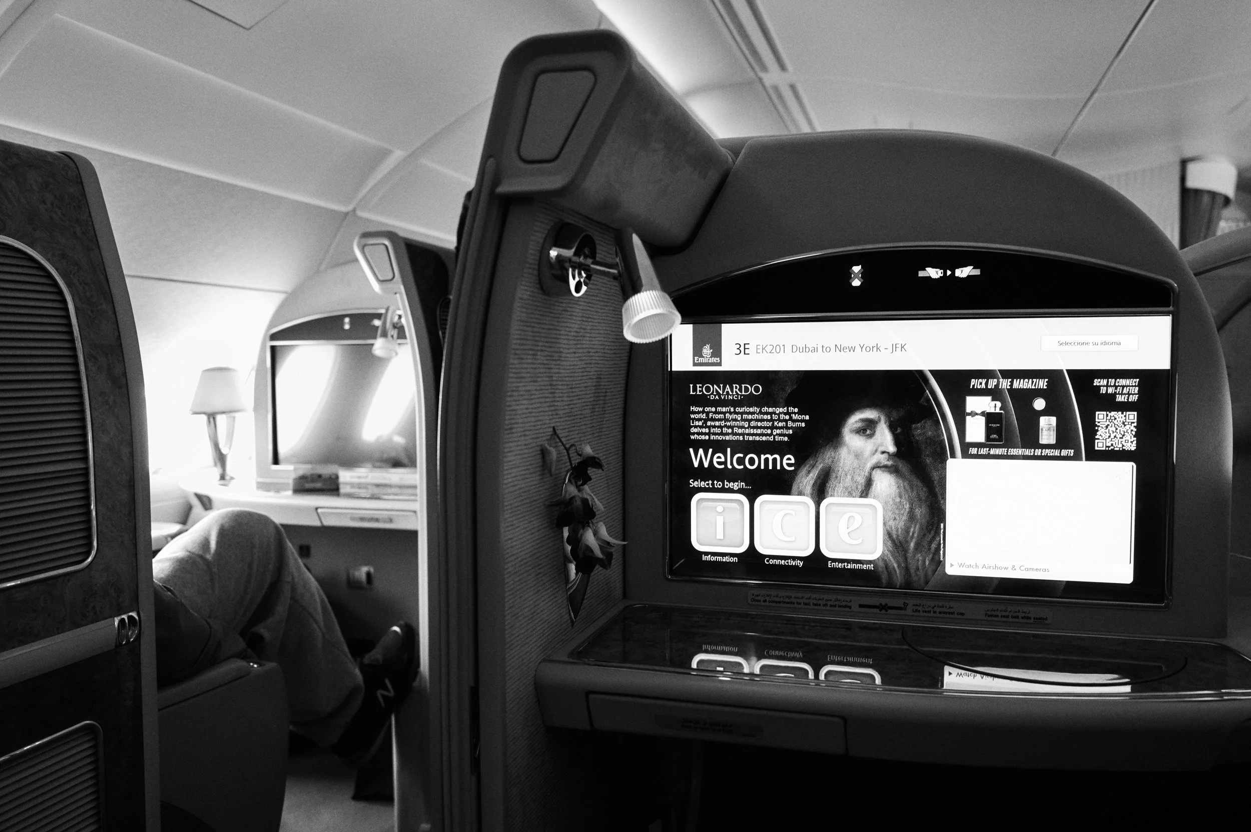 In-flight entertainment screen showing a welcome message and options for information, connectivity, and entertainment on a Emirates flight from Dubai to New York, with a man seated in front.