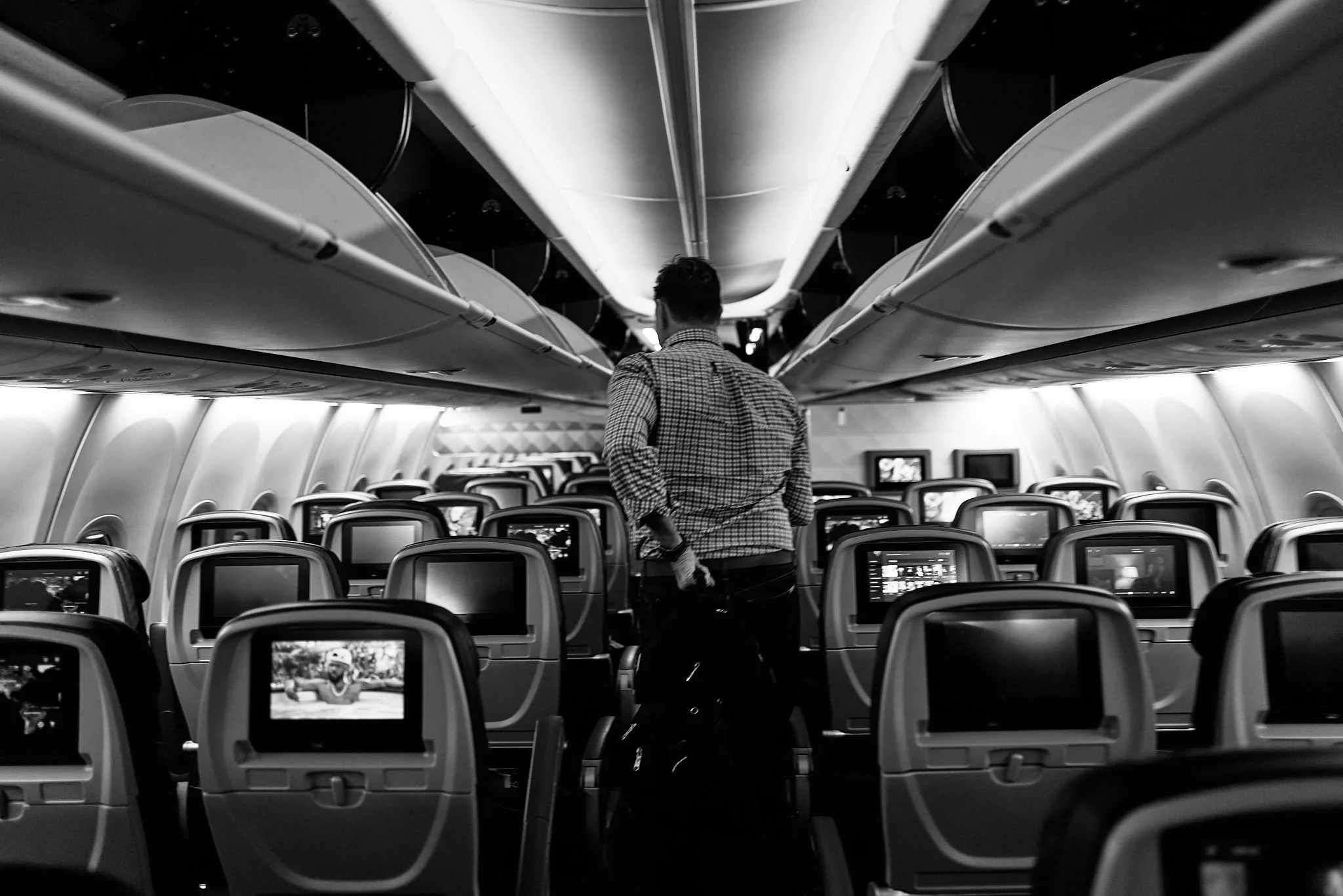Black and white photo of a man with a backpack walking down the aisle of an airplane cabin between rows of seats with in-flight entertainment screens  by Matt Ellis