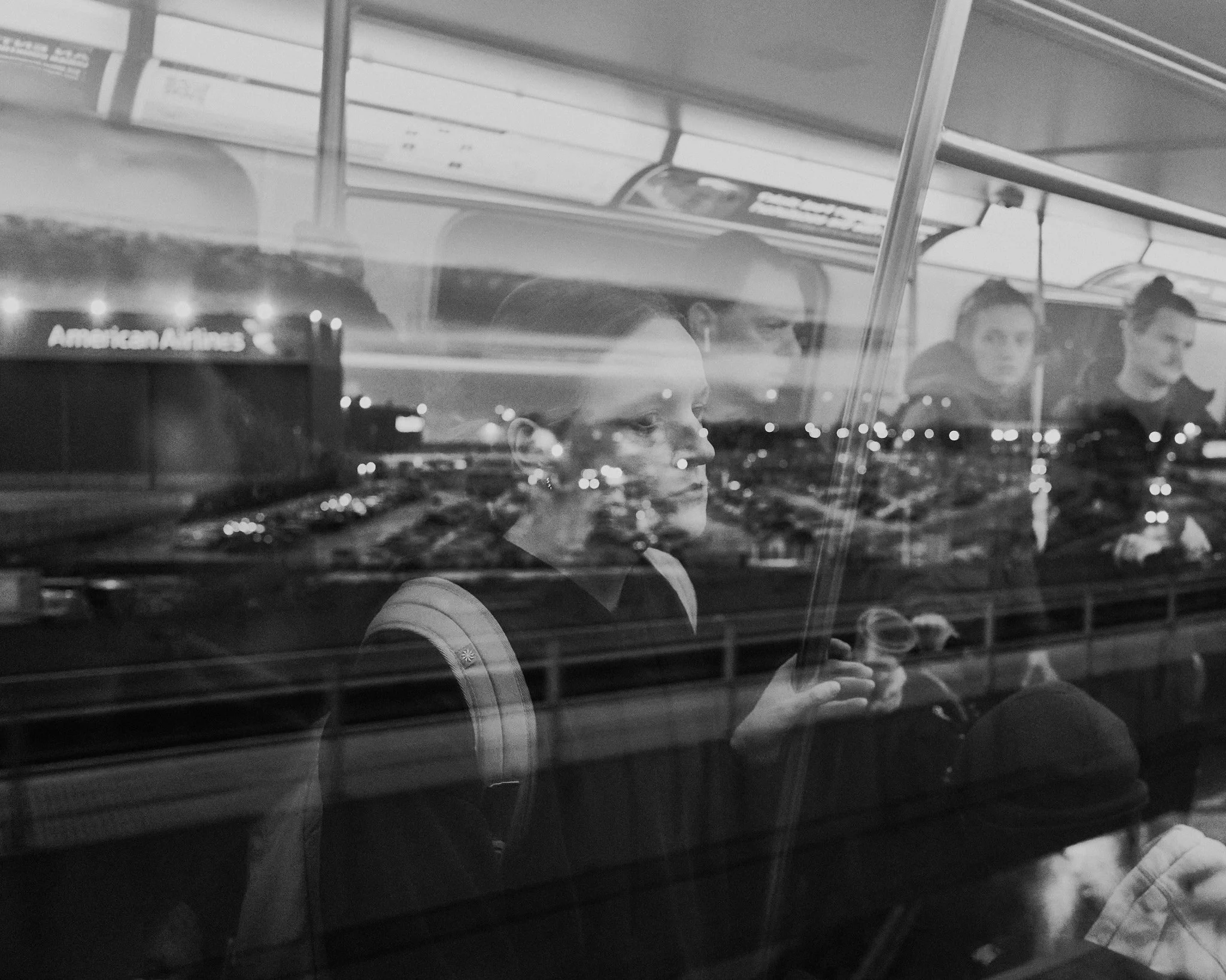 Black and white photo of several people inside a moving train, view of the city and an airport in the background through the train window at night.