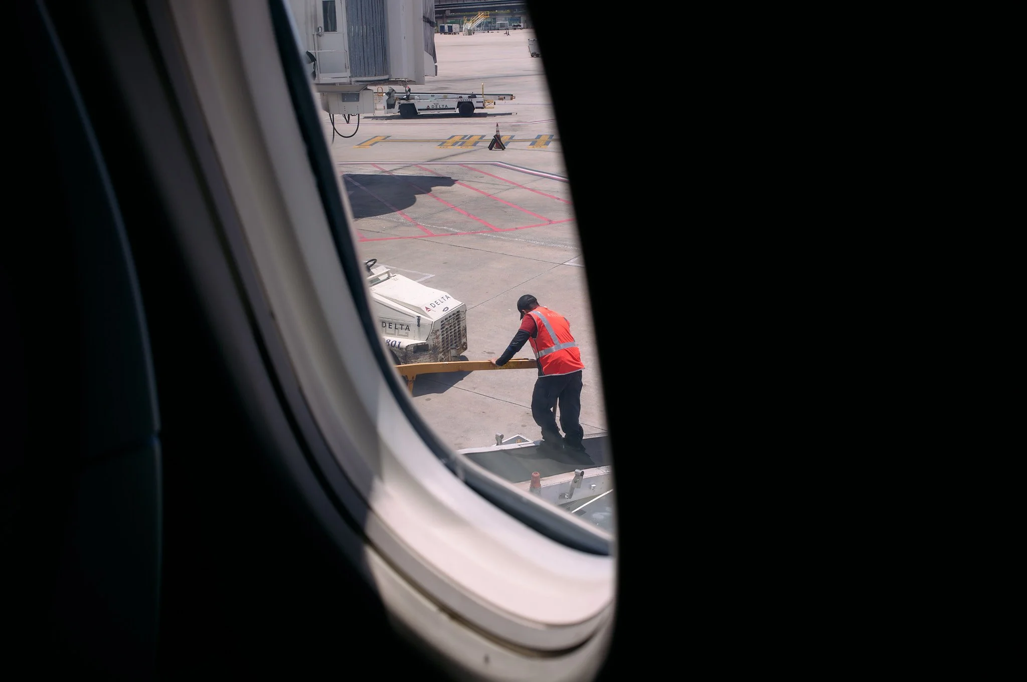 A worker in a red safety vest and a black cap is pushing a yellow luggage cart on the tarmac at an airport, viewed through an airplane window  by Matt Ellis