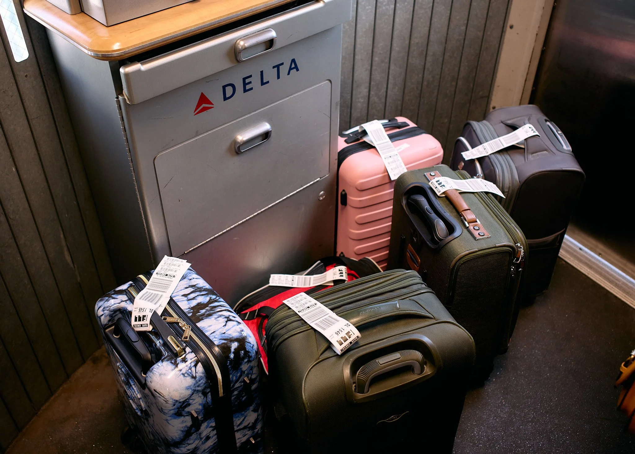 Five pieces of luggage, including a blue and white patterned suitcase, a pink suitcase, a dark green suitcase with a brown strap, a black suitcase, and a black bag, are placed on the floor against a metal security box with the DELTA logo in an airpor
