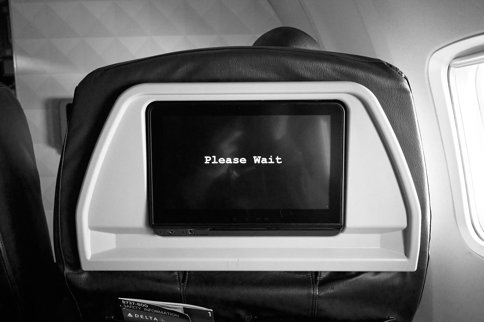 In-flight entertainment screen on the back of an airplane seat displaying 'Please Wait' in white text.  by Matt Ellis