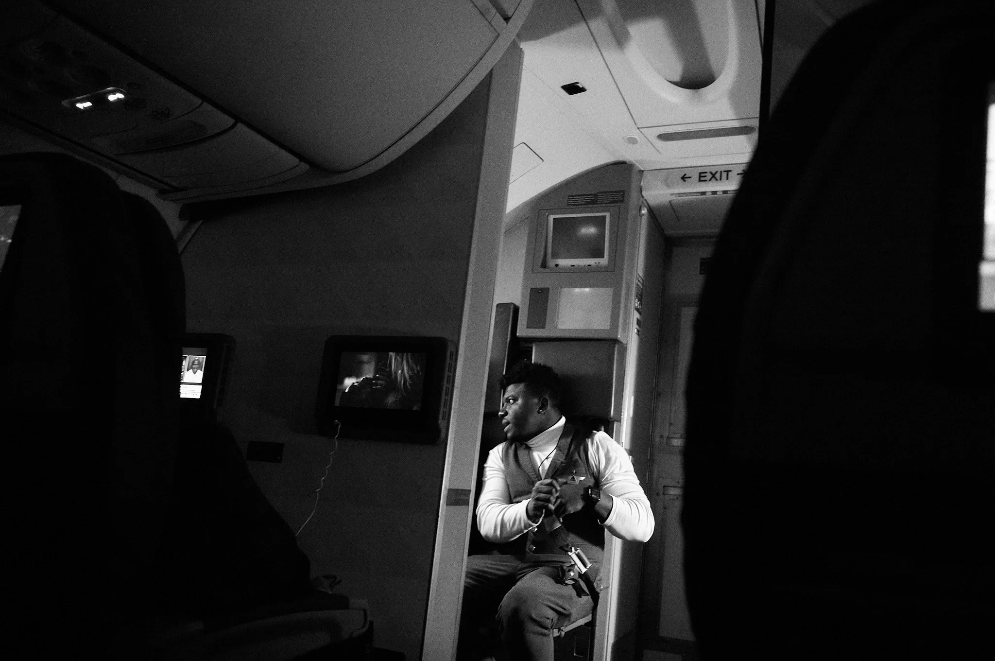 A man sitting in a seat on an airplane, facing sideways, wearing a white shirt and a vest, with headphones around his neck. The airplane interior is visible, including overhead compartments, monitor screens, and an 'EXIT' sign.