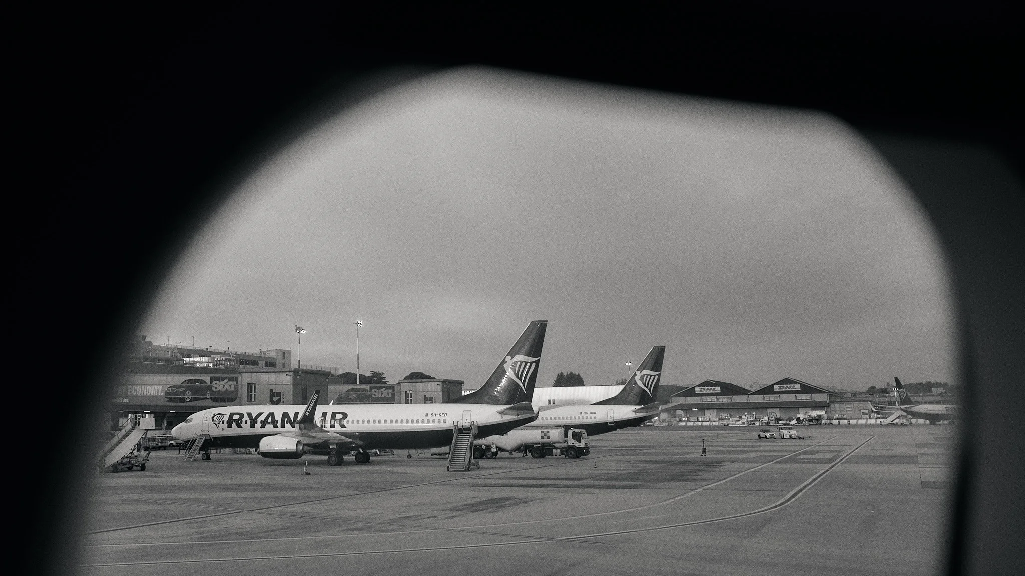 View of an airport tarmac with two Ryanair airplanes parked, seen through an airplane window  by Matt Ellis