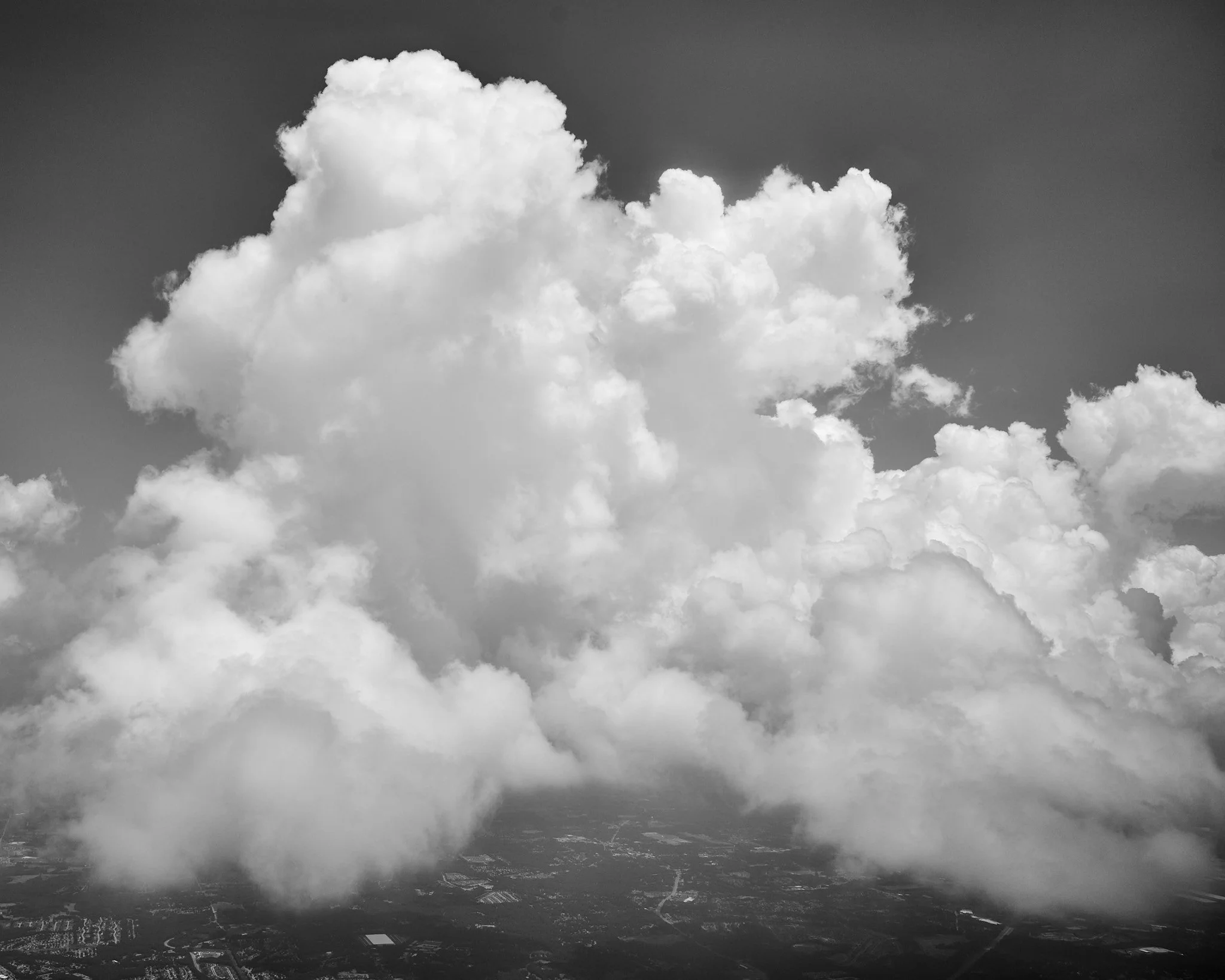 Black and white photo of large, fluffy clouds in the sky over a landscape with roads and fields below.