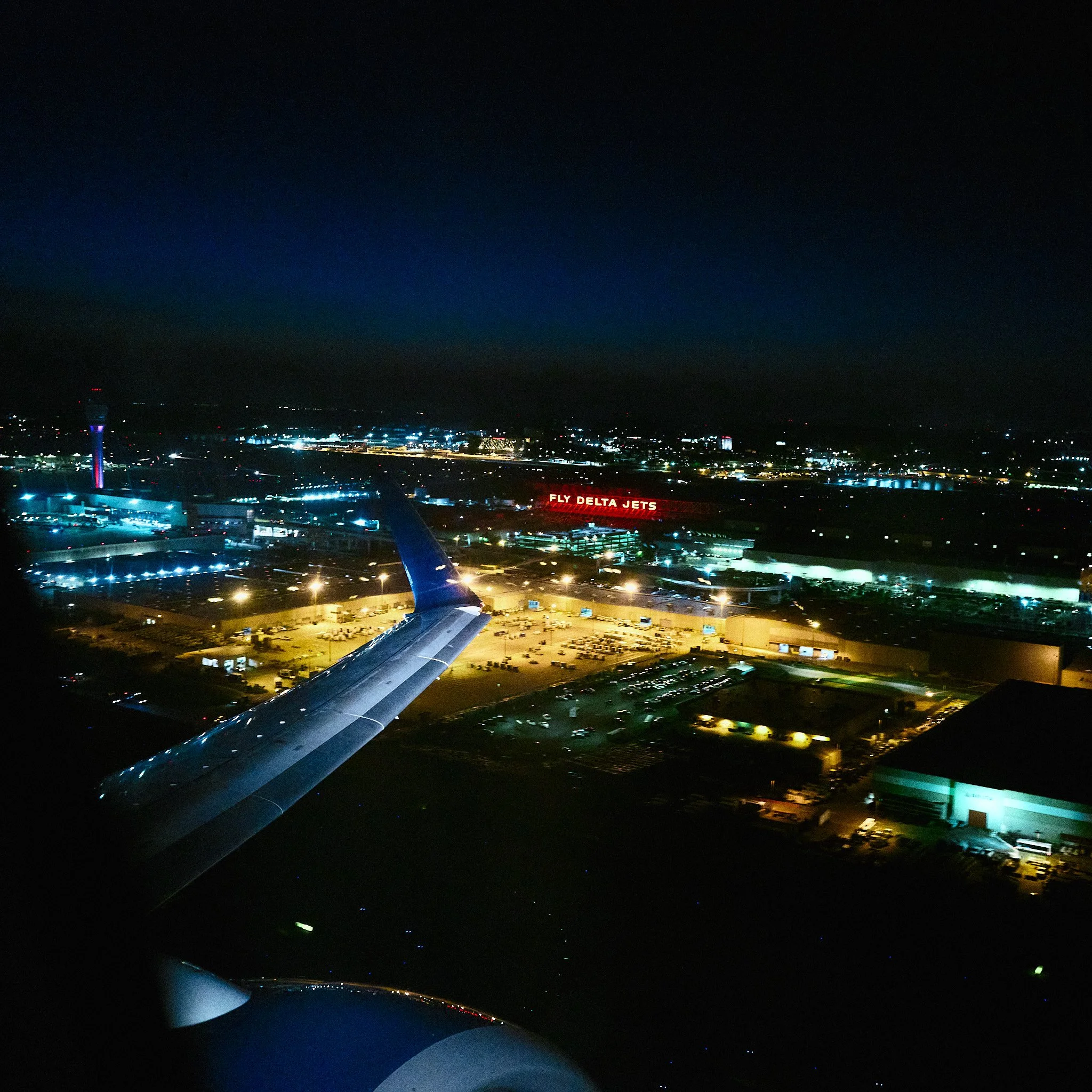 Nighttime view from an airplane window showing the airplane wing and illuminated airport and city lights below, with a sign reading 'Fly Delta Jets' in the distance at Hartsfield ATL airport