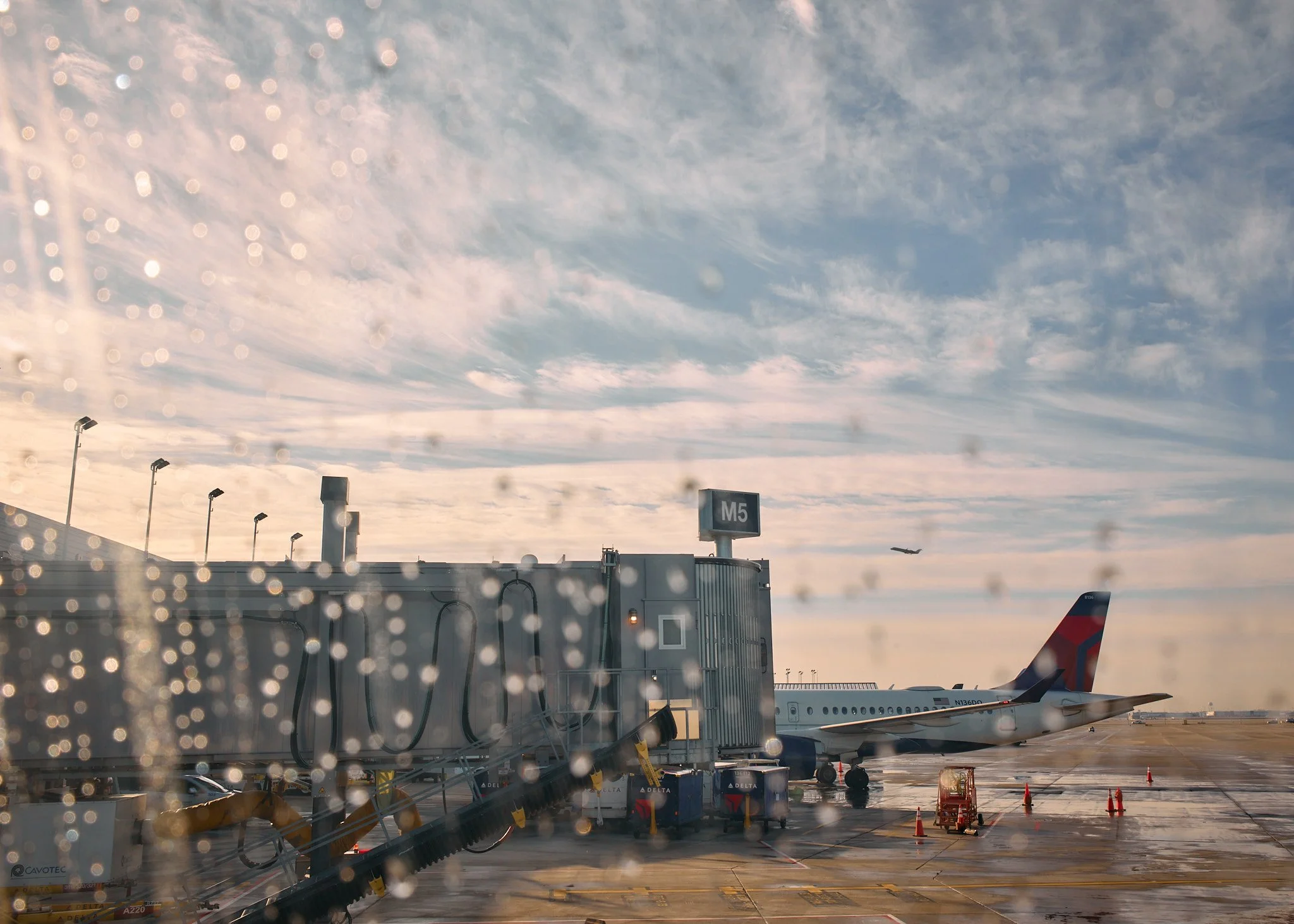 View of an airport tarmac through a rain-speckled window, showing a parked airplane, jet bridge, and a plane taking off in the sky with clouds  by Matt Ellis