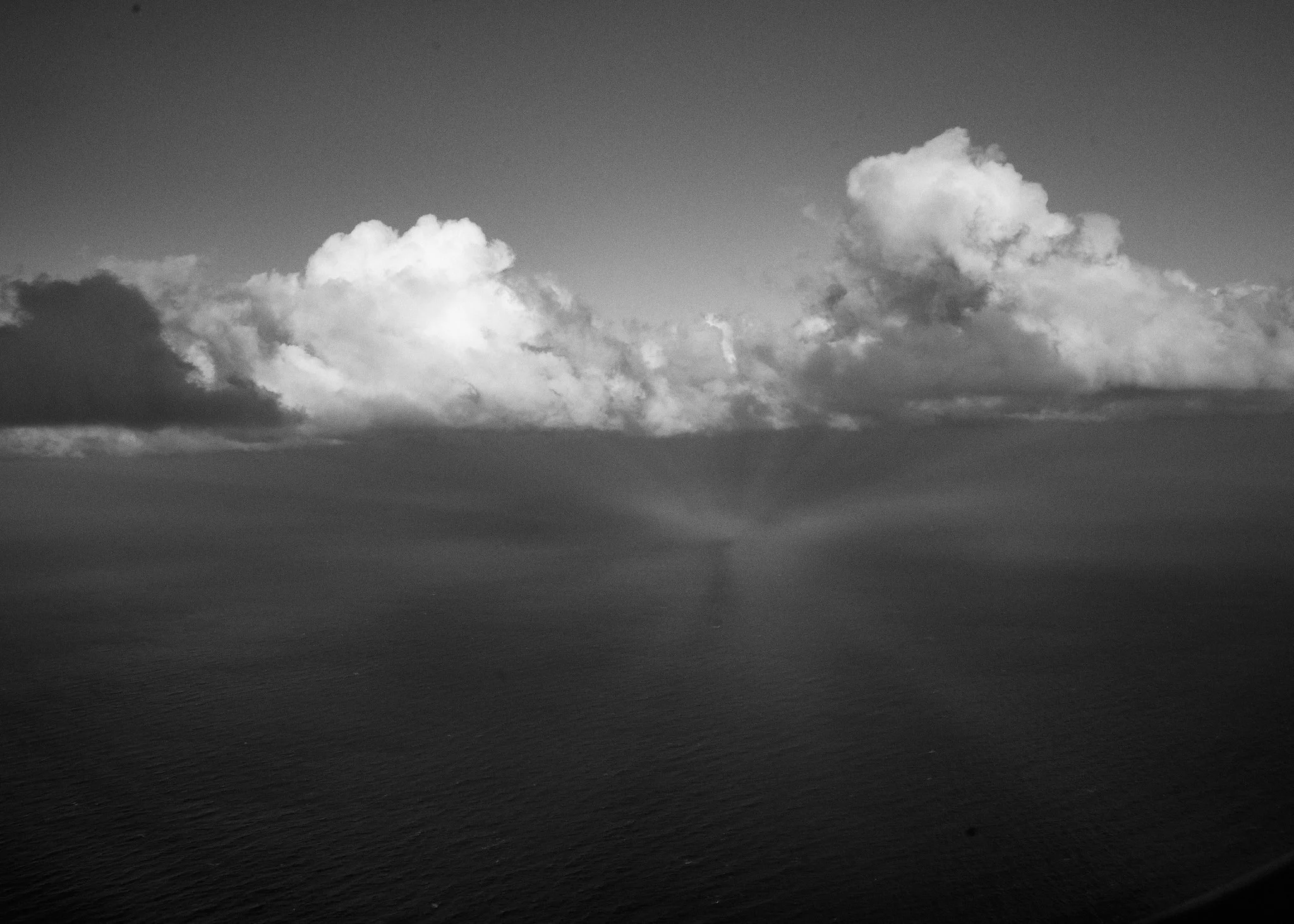 Black and white photo of the ocean with a large, fluffy cloud above and sun rays breaking through the clouds.