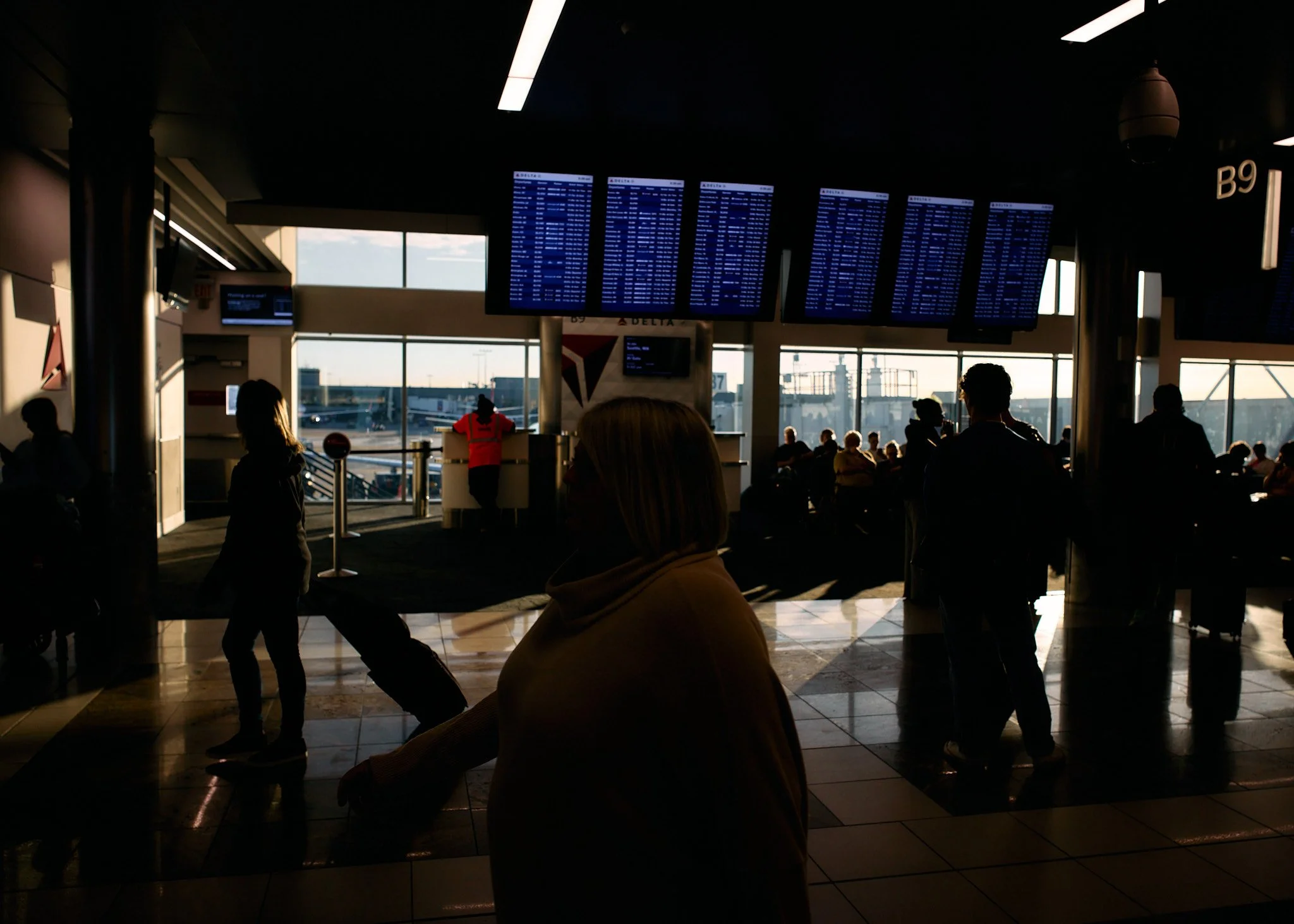 Passengers waiting at an airport terminal with flight information displays overhead, some silhouettes in front of large windows showing the tarmac and planes outside.