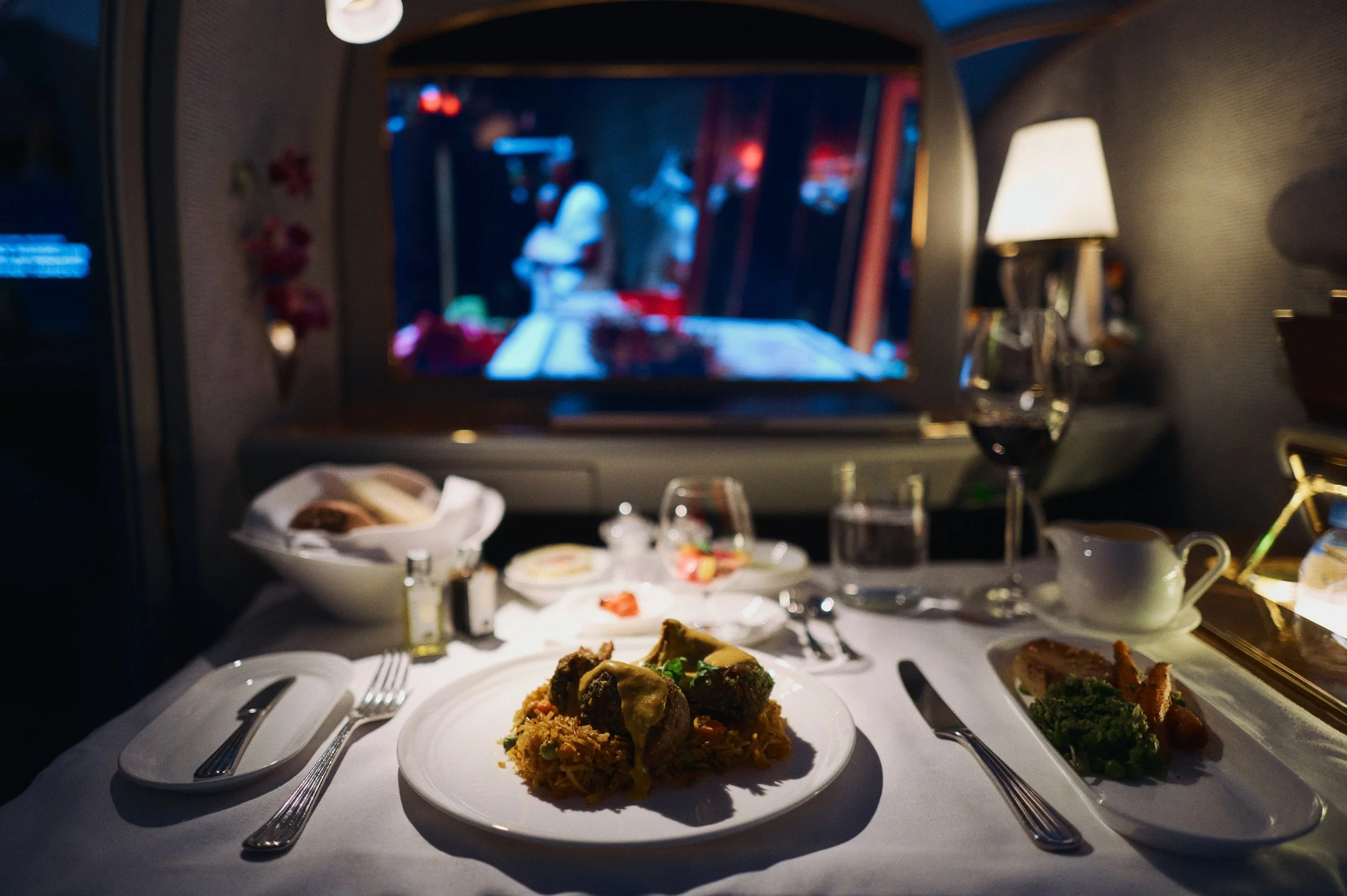A meal served on a white tablecloth inside a private jet, featuring a plate of meat and rice, a side dish with vegetables, a bowl of bread, a glass of red wine, a cup and saucer, with a window showing a blurred outdoor scene.