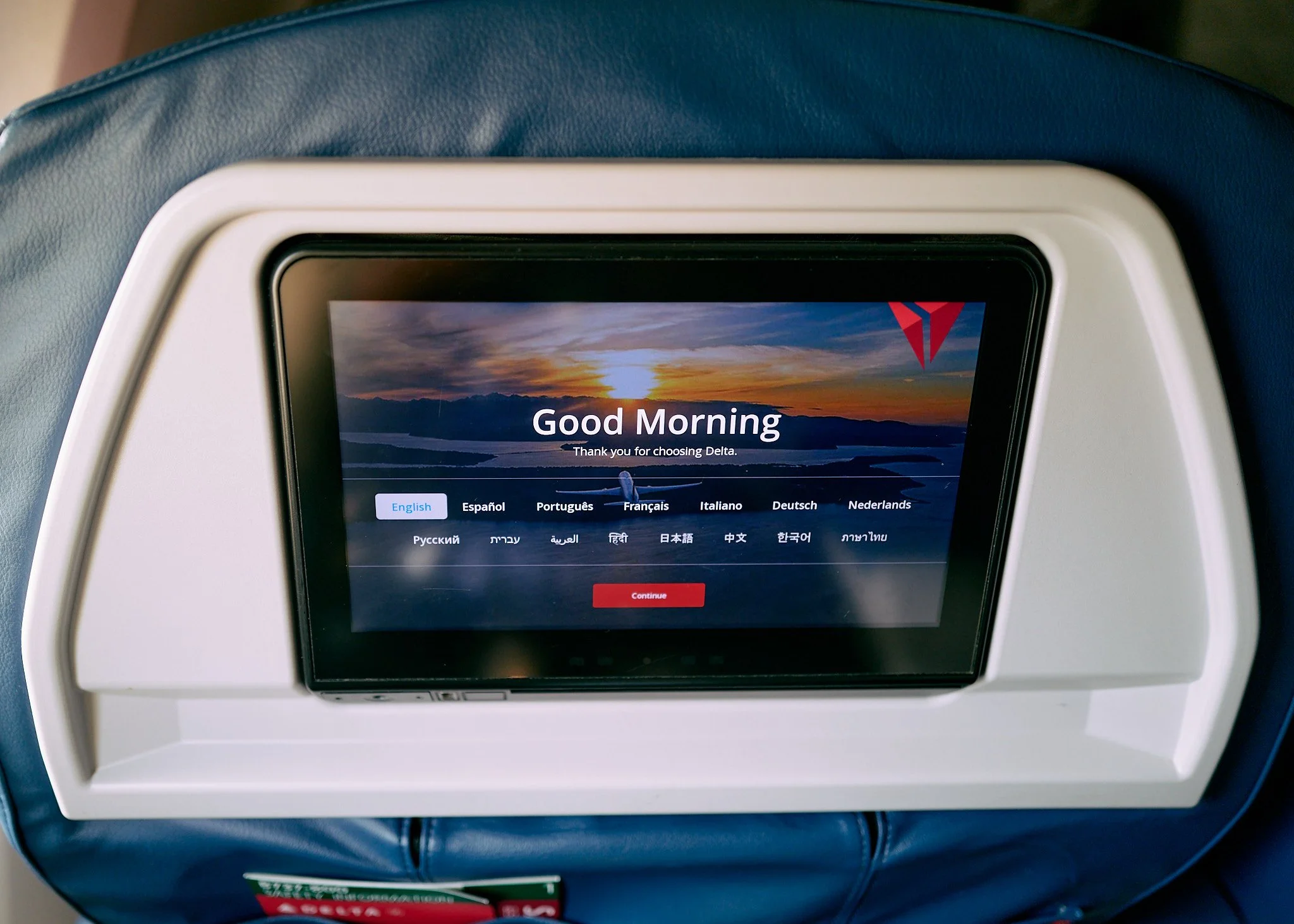 In-flight entertainment screen displaying a sunset background, Delta Airlines logo, and a greeting 'Good Morning' in multiple languages with a 'Continue' button, integrated into the seat back of an airplane  by Matt Ellis