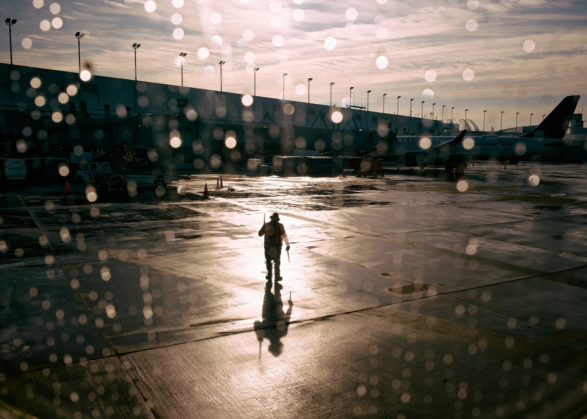 An airport tarmac scene at sunset or sunrise, with rain droplets on the window in the foreground. A person with a backpack is walking away from the camera towards airplanes parked at gates, with the wet ground reflecting the sunlight.