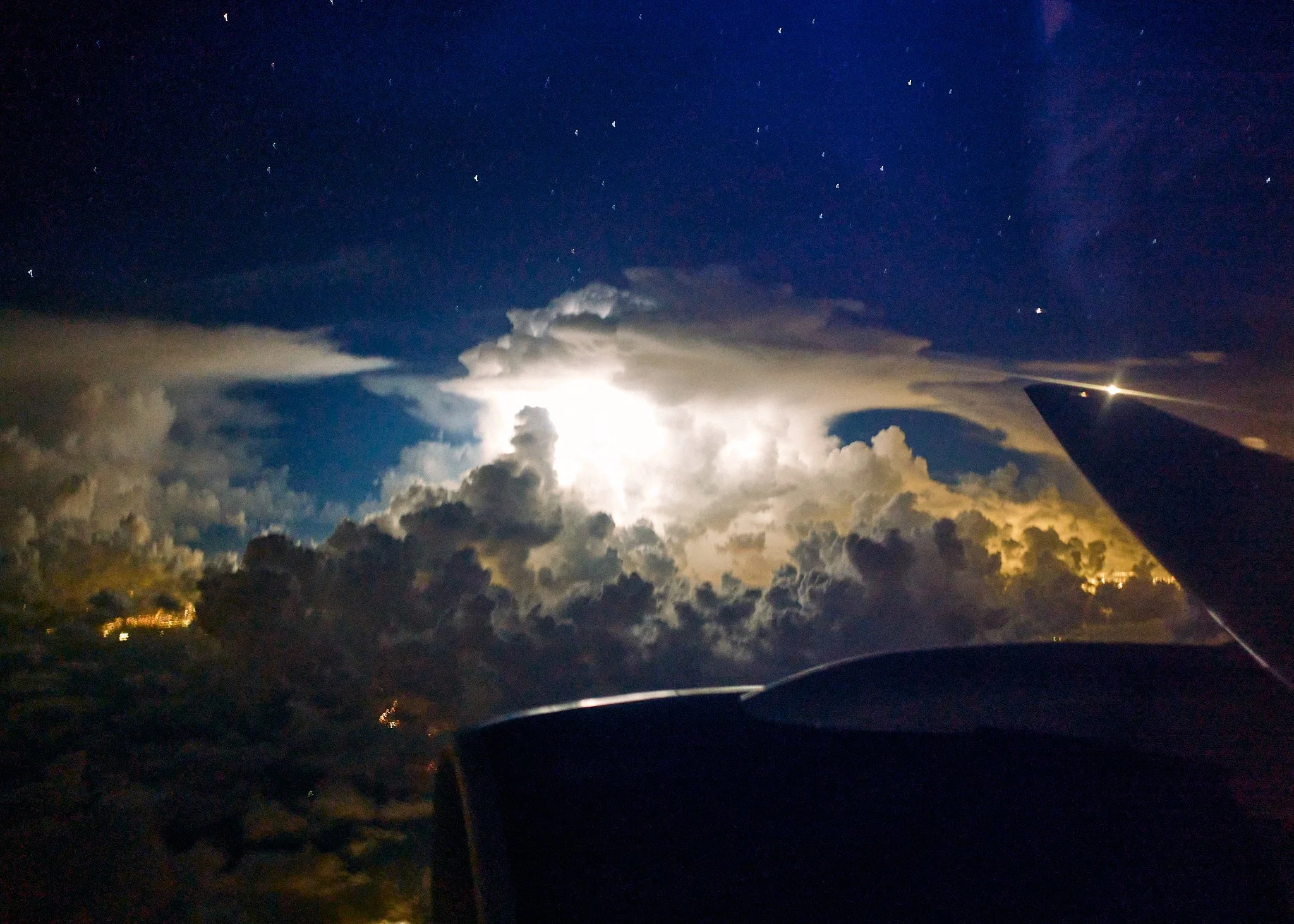 Night sky view from an airplane window, with clouds illuminated by lightning and stars visible in the background, airplane wing visible on the right side