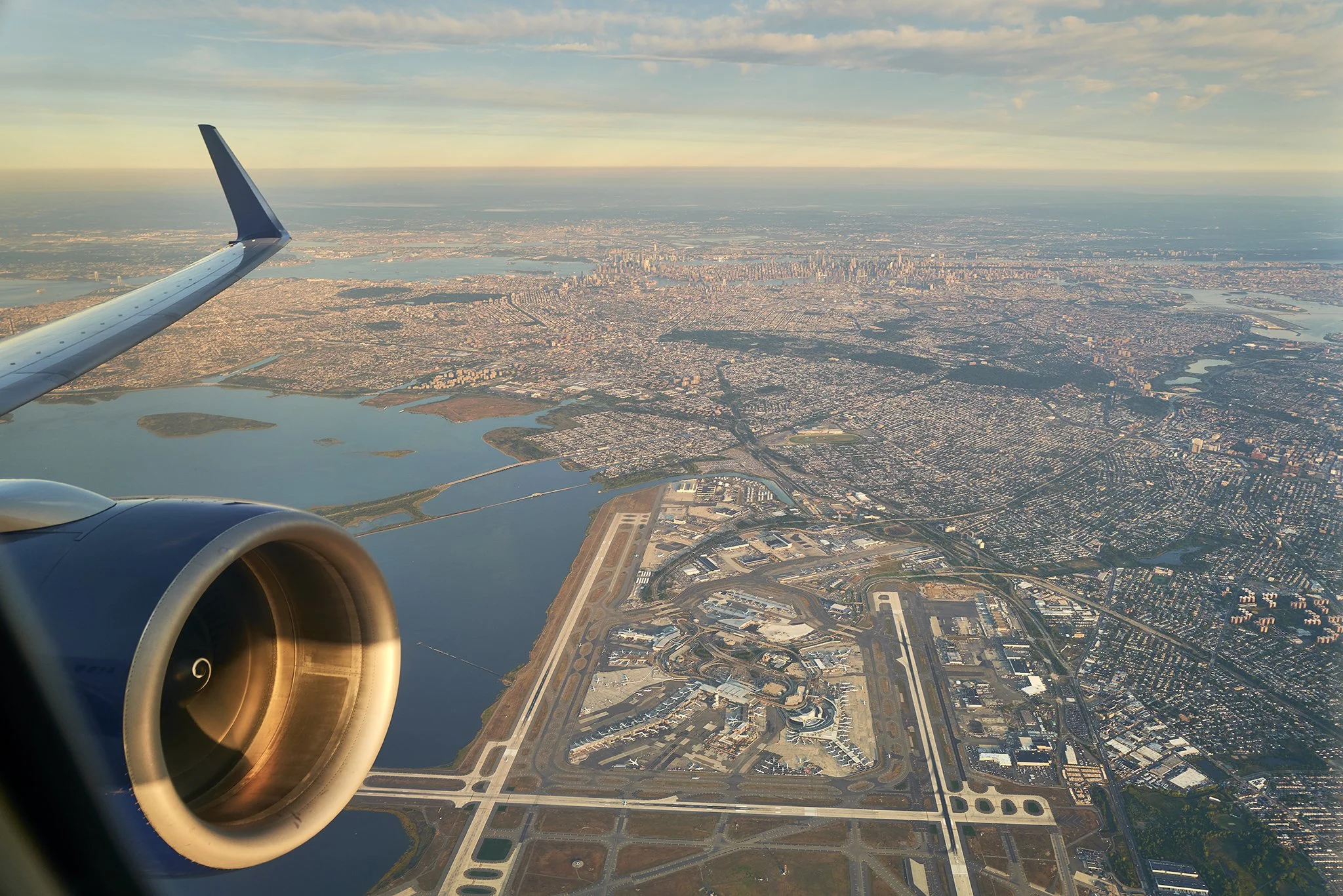 Aerial view from an airplane window showing a cityscape with water bodies, highways, and a runway, during sunset.