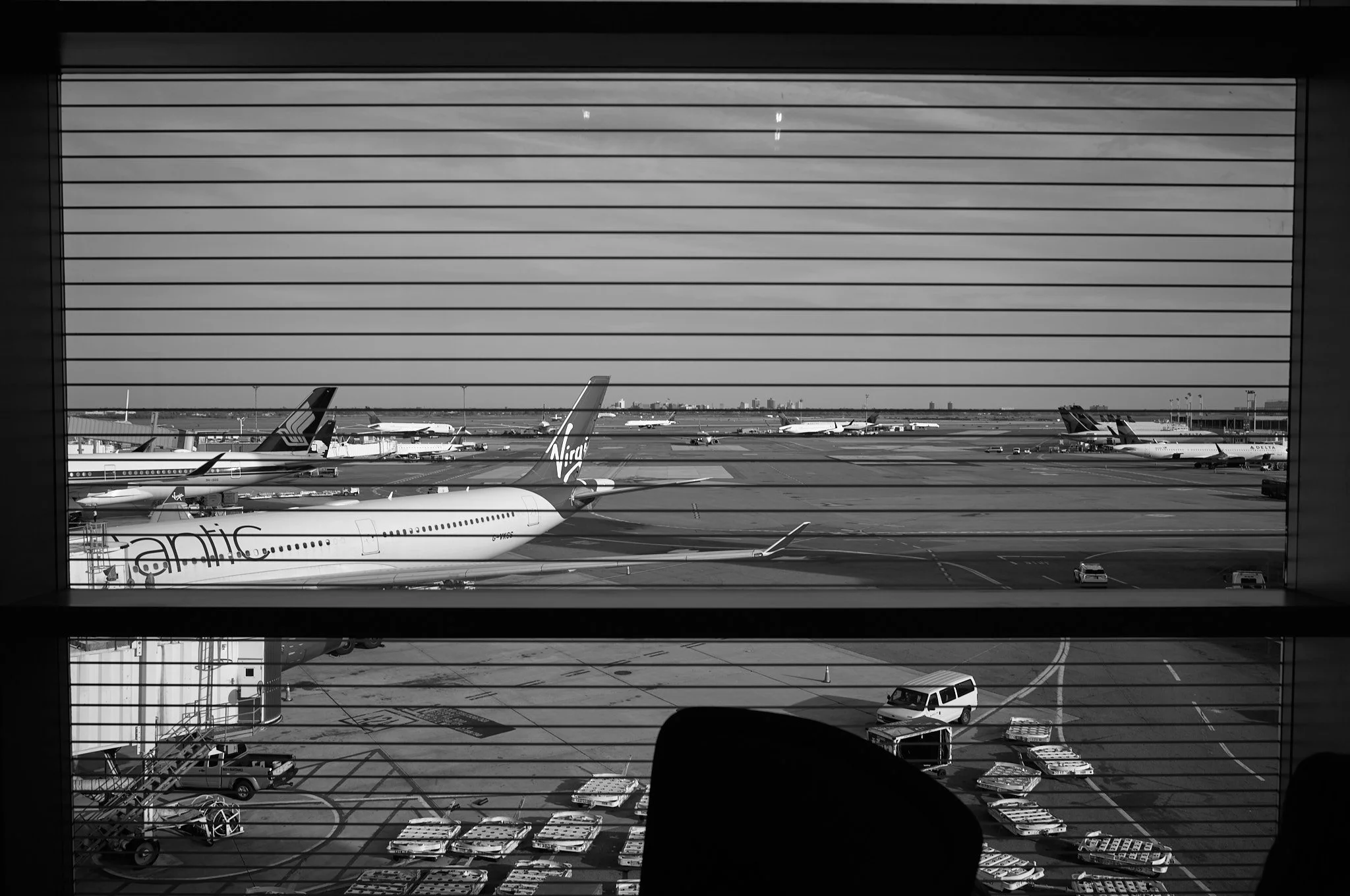 View of jfk airport tarmac through window blinds, with airplanes parked and luggage carts on the ground.
