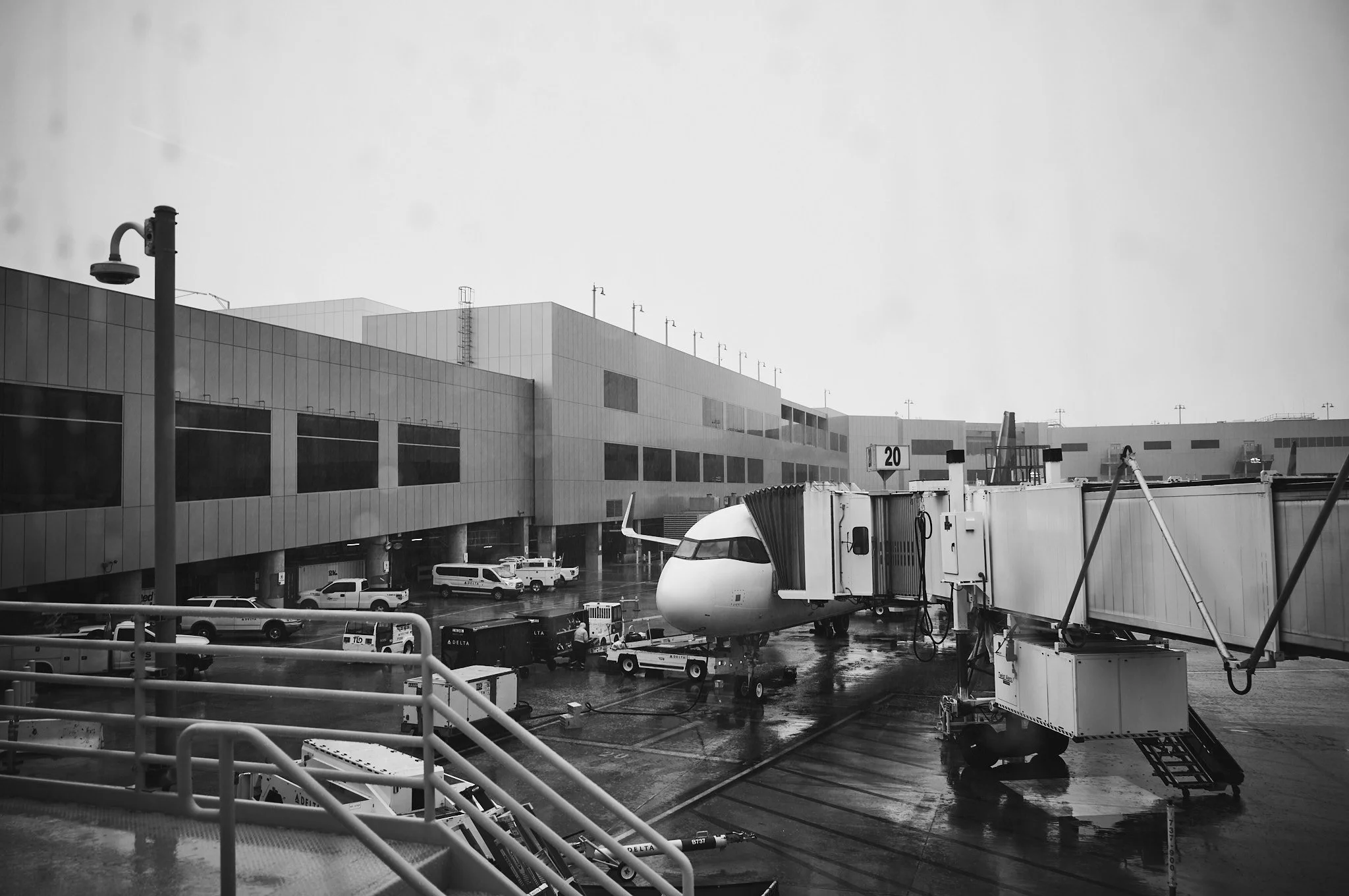 A black and white photograph of an airport terminal with a parked airplane connected to a jet bridge, surrounded by baggage carts, vehicles, and airport infrastructure on a wet tarmac  by Matt Ellis