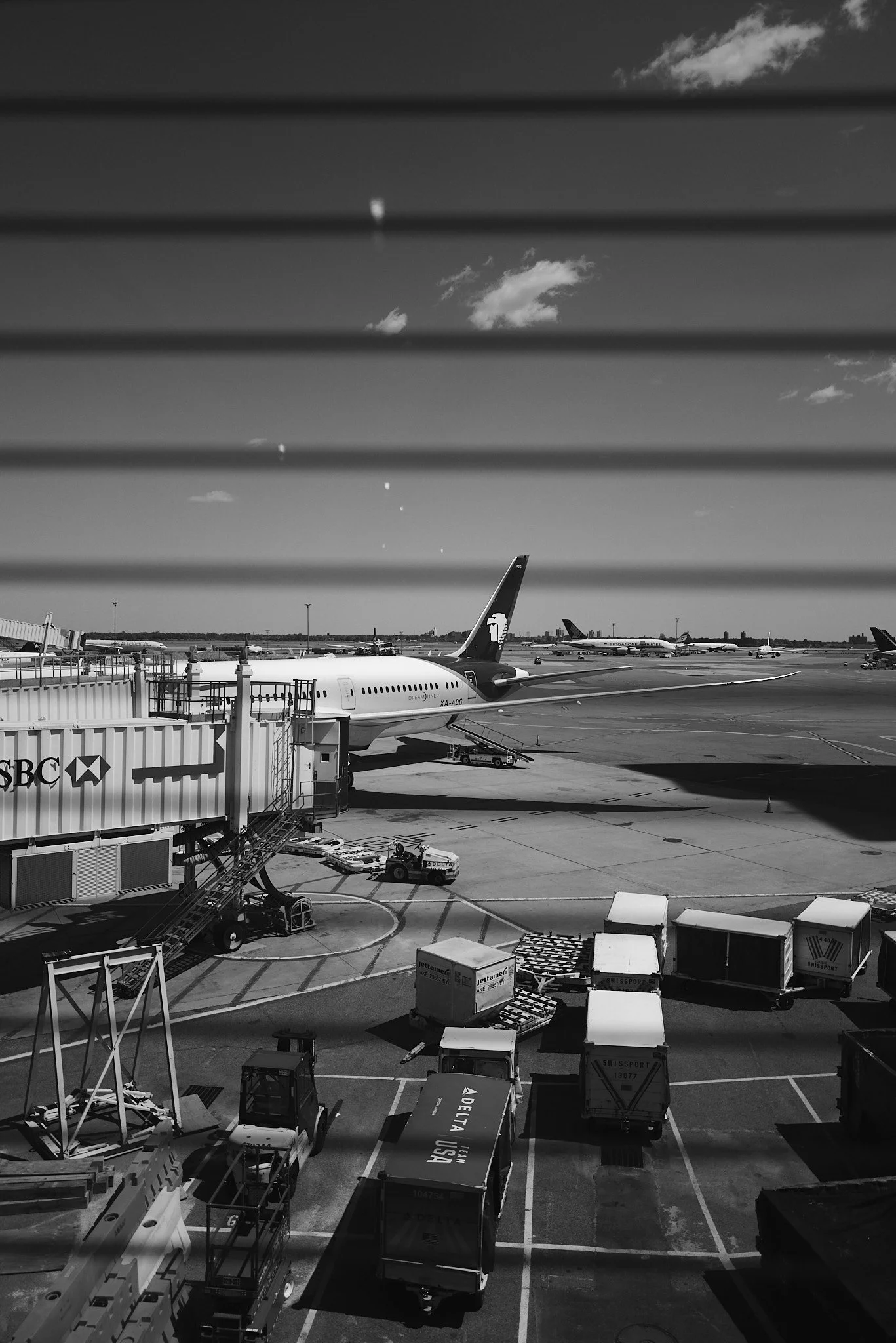 Black and white photo of jfk airport tarmac with airplanes parked at gates and luggage carts in the foreground.
