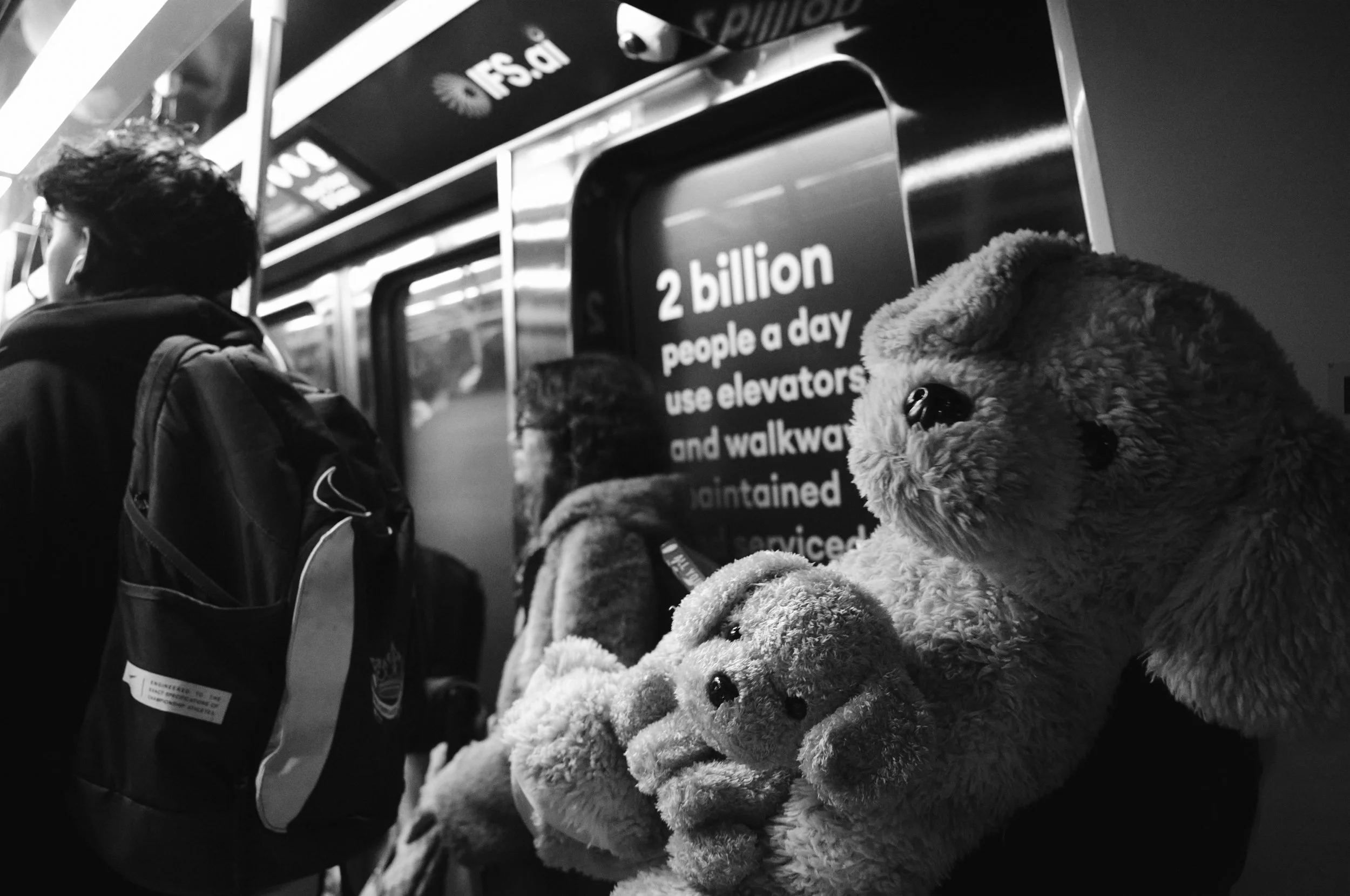 People standing in line on a subway platform, with a person holding two teddy bears in the foreground.