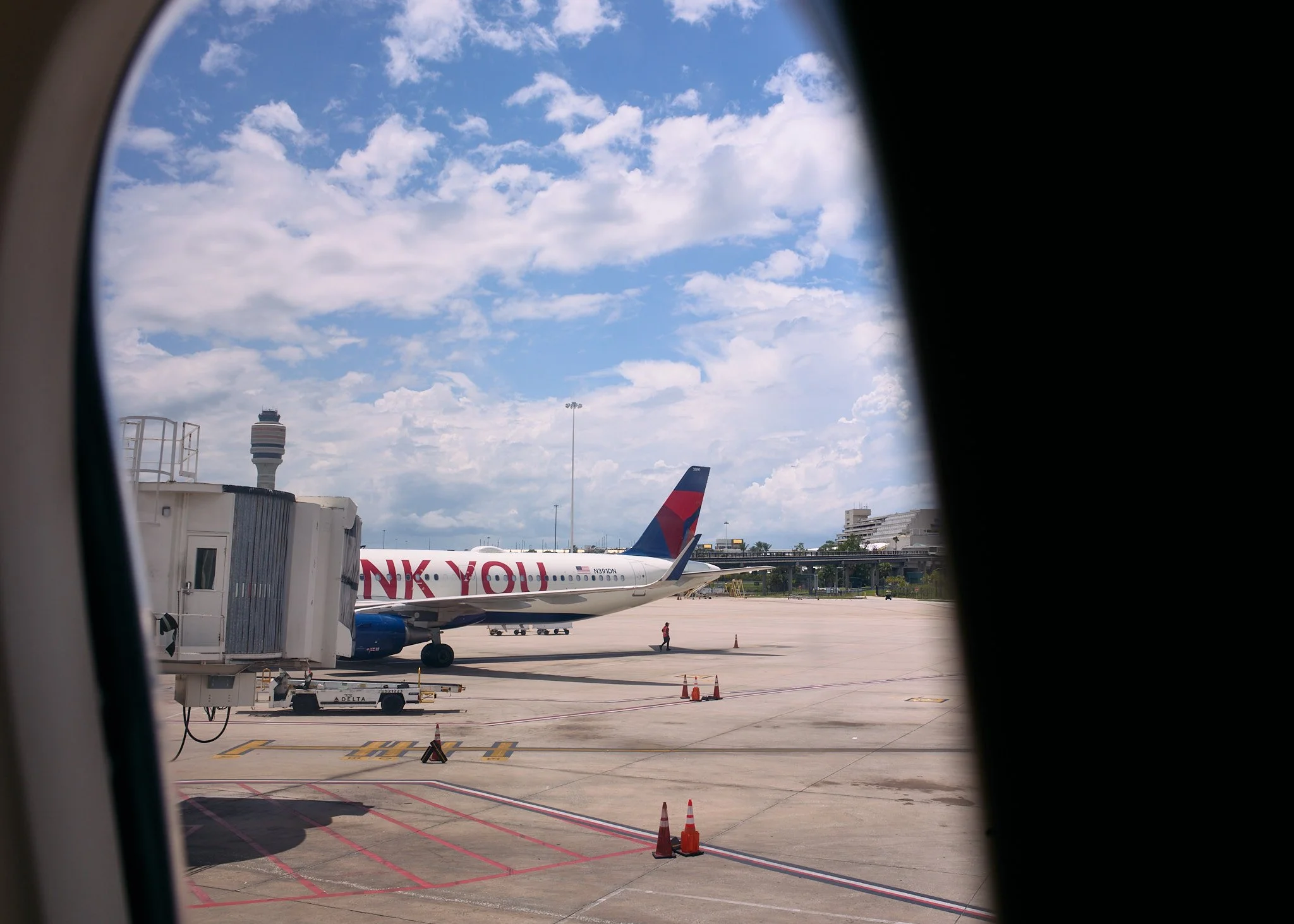 View of an airport tarmac seen through an airplane window, featuring a Delta airplane with 'THANK YOU' written on the side, airport buildings, and a control tower in the background  by Matt Ellis