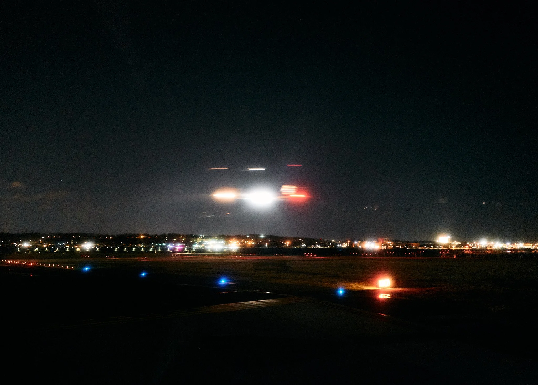 Nighttime airport runway with illuminated blue and red lights and a distant city skyline in the background. The sky is dark with some faint stars  by Matt Ellis