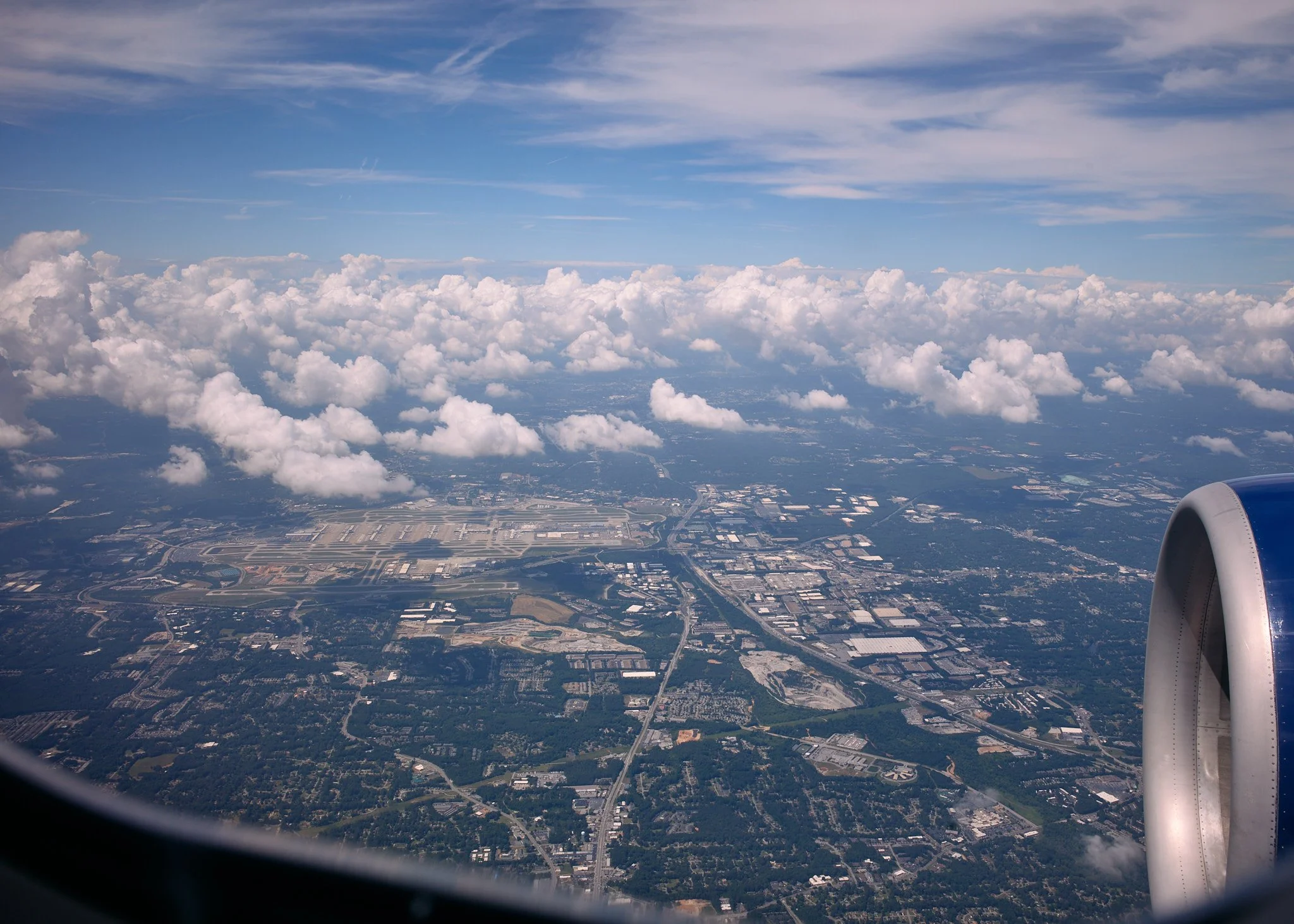 Aerial view from airplane window showing clouds and a cityscape below, with part of the airplane engine visible on the right  by Matt Ellis