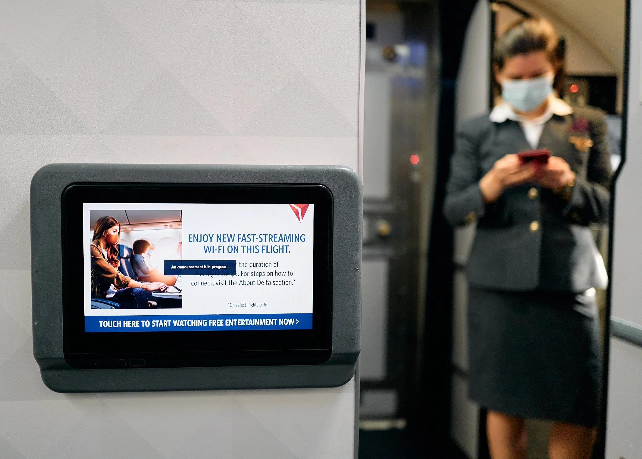 In-flight screen displaying advertisement for Wi-Fi service with a photo of a woman wearing a mask and using a laptop on an airplane, in the background a flight attendant in uniform with a face mask is standing near on the phone  by Matt Ellis