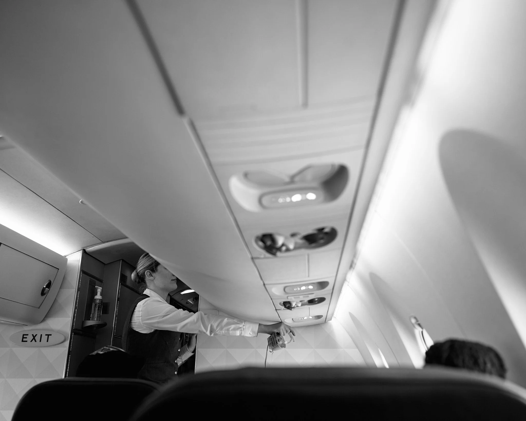 An airline flight attendant standing in the aisle of a plane, reaching towards the overhead storage compartment with a drink in her hand. Passengers are seated below, and an exit sign is visible  by Matt Ellis