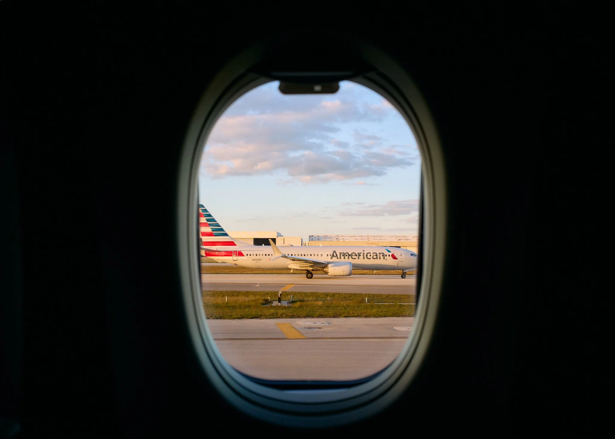 An airplane with American Airlines livery, viewed through an airplane window, parked on the tarmac at an airport during sunset.