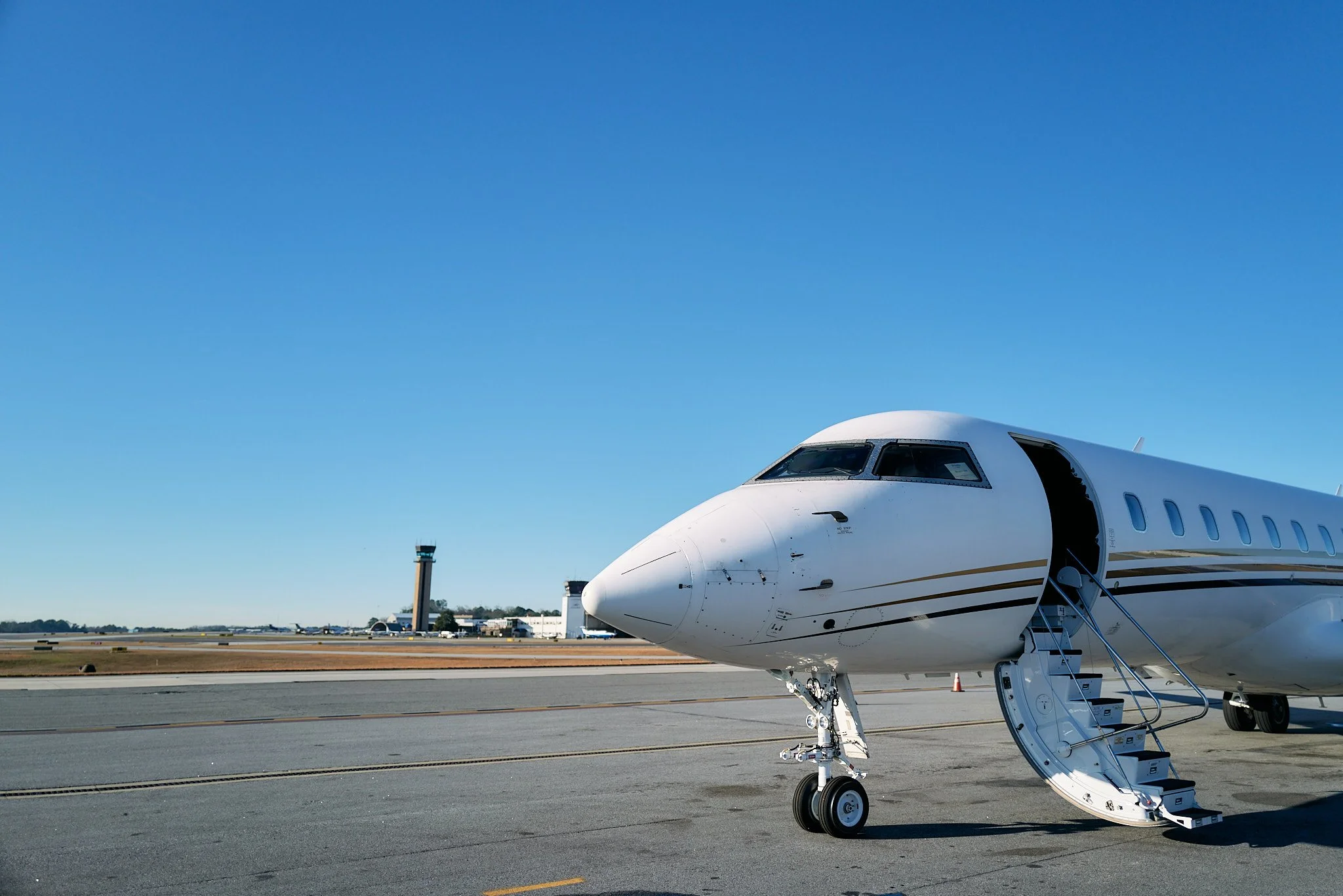 A private jet parked on an airport tarmac with its door open and stairs extended. Airport control towers and hangars in the background under a clear Blue sky.