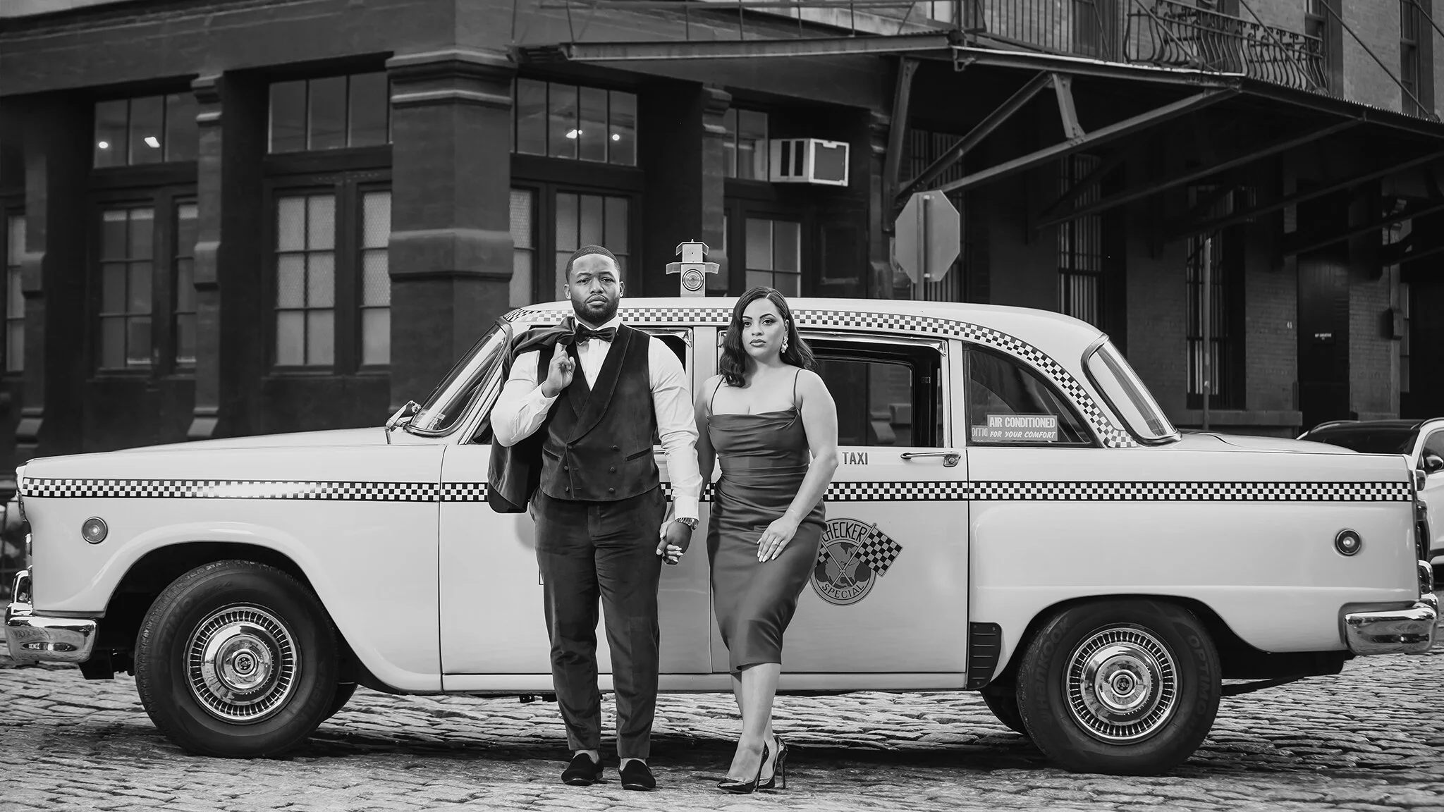 A man and woman in formal attire standing in front of a vintage taxi car on a cobblestone street.