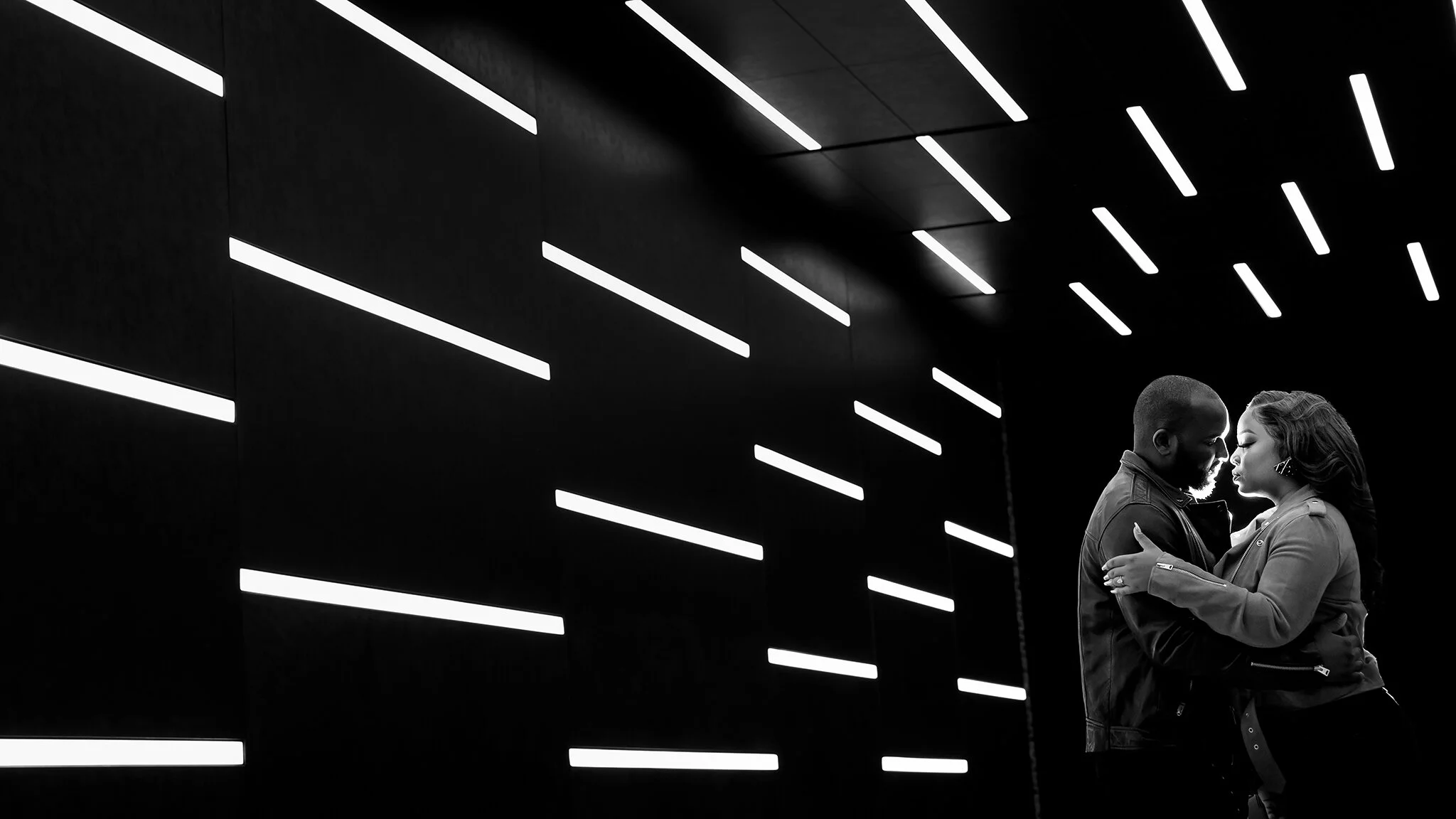 A black and white photo of a couple embracing against a black wall with bright horizontal and diagonal white light strips.