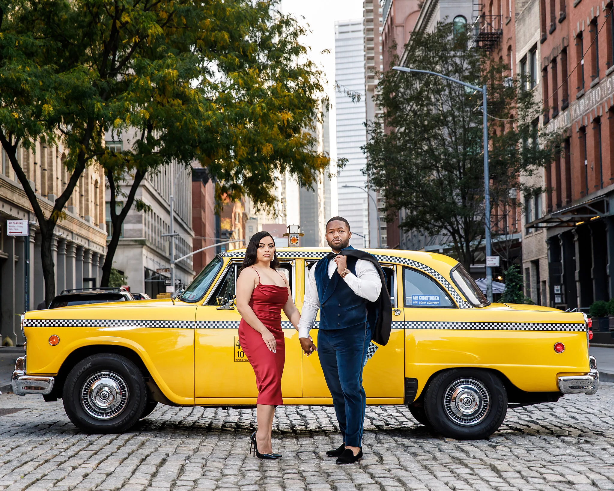 A man and woman dressed formally standing hand in hand on a cobblestone street in front of a yellow taxi cab in an urban area with buildings and trees.