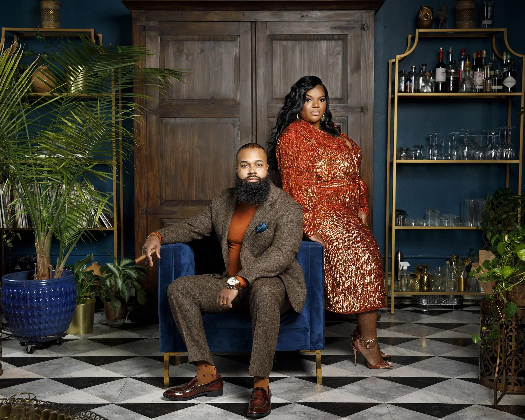 A stylish man with a beard in a brown suit and orange shirt sitting on a blue velvet armchair, with a woman in a sparkly orange dress standing behind him in a room with blue walls, wooden door, and glass shelves with bottles and glasses, surrounded b