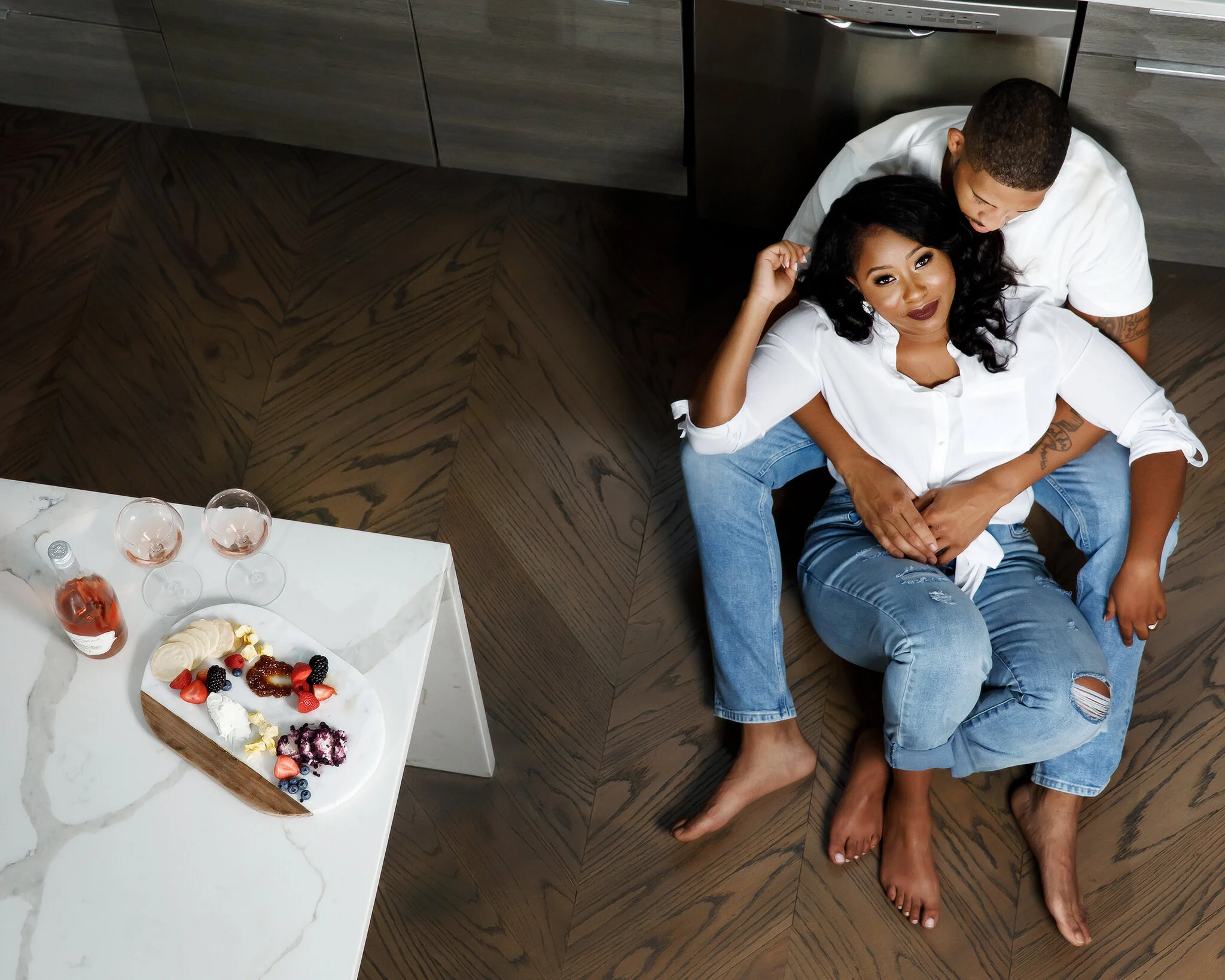 A woman and man sitting closely together on the floor, with the woman leaning against the man. They are both sharing a relaxed moment in a modern kitchen, with the woman looking at the camera. There is a marble table nearby with a dessert platter, st