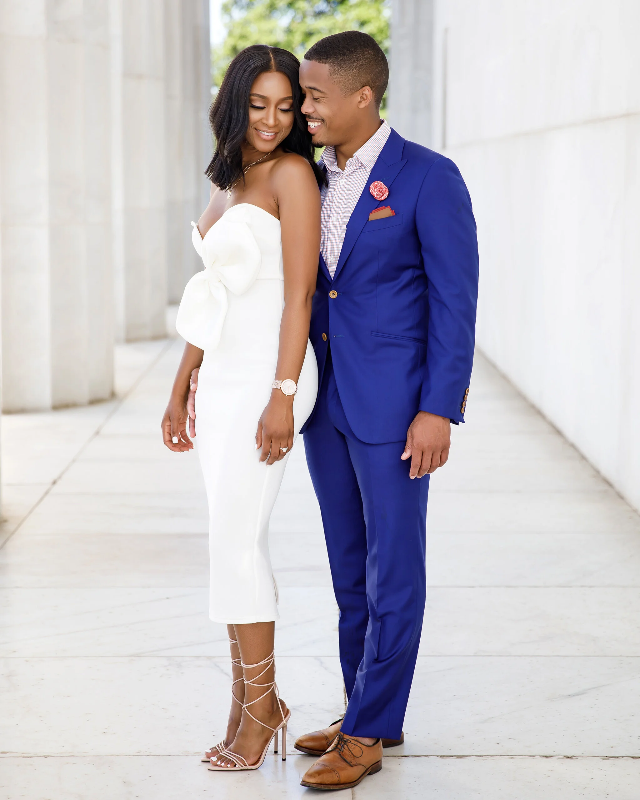 A couple dressed in formal attire standing close together, smiling and looking at each other in an outdoor setting with white columns and greenery in the background.