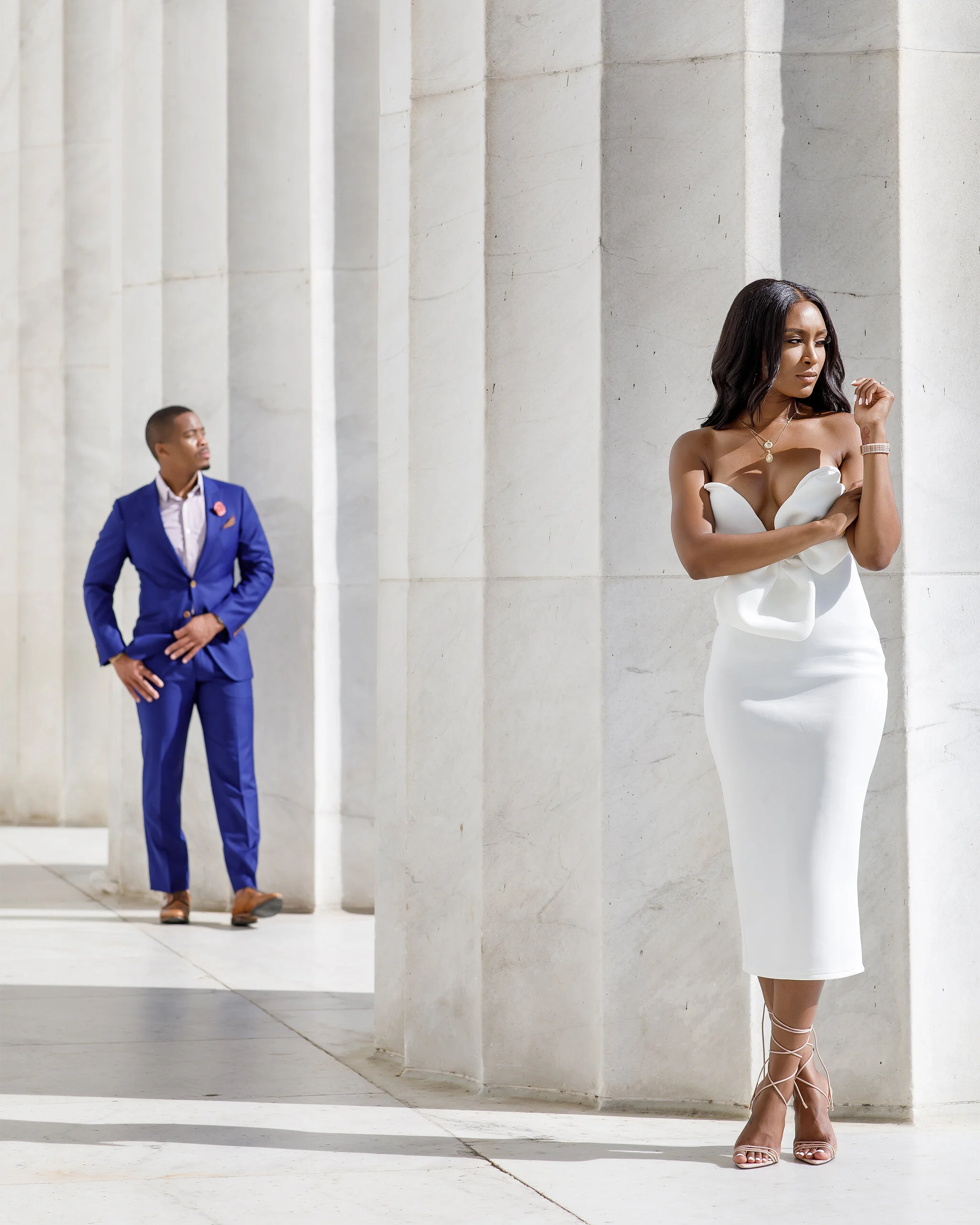 A woman in a white dress stands alone in the foreground, while a man in a blue suit stands at a distance behind her, both positioned near large stone columns.