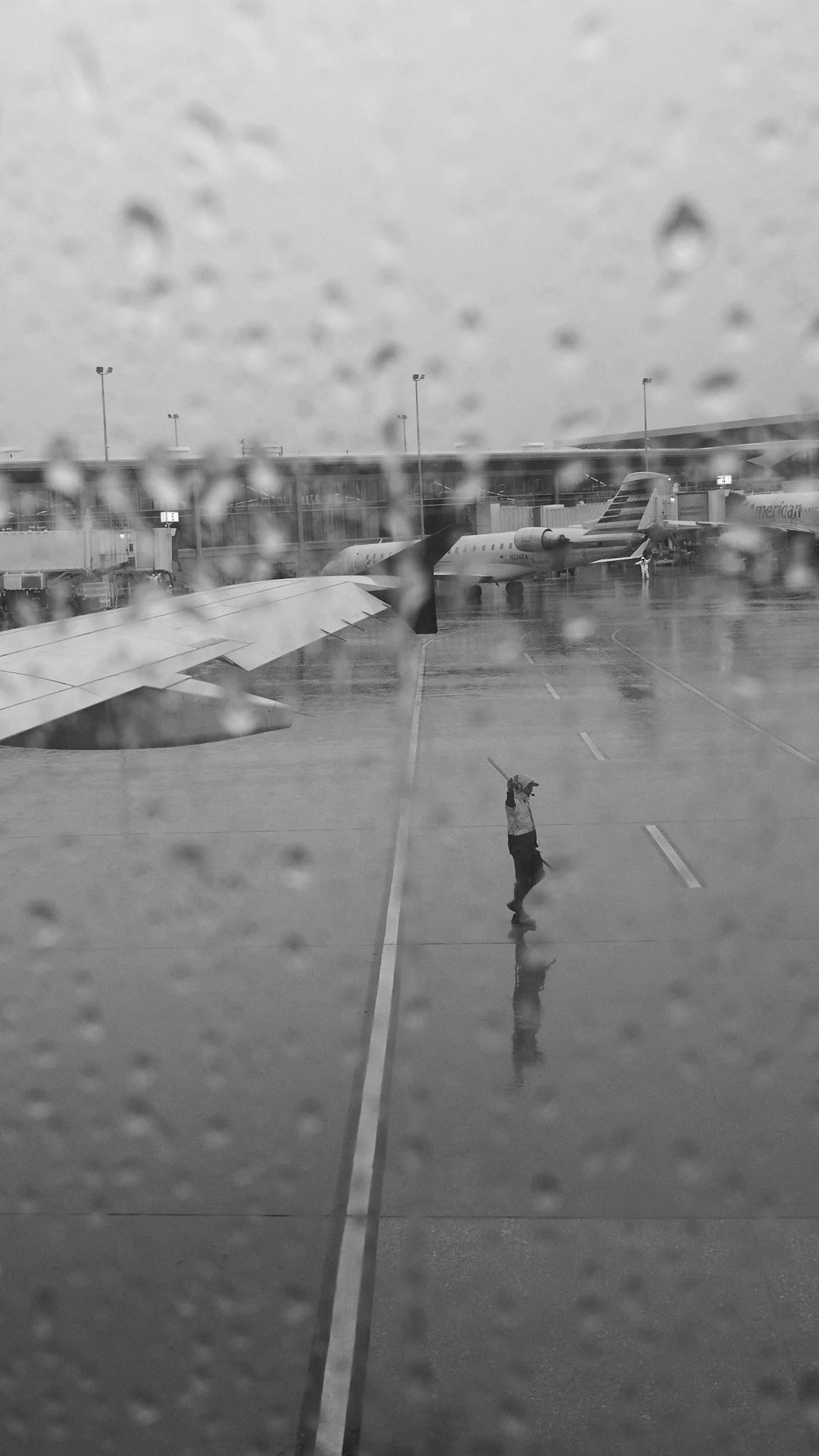 Rainy day at an airport seen through a window with raindrops; an aircraft wing and a ground crew member holding a stick are visible on the tarmac.