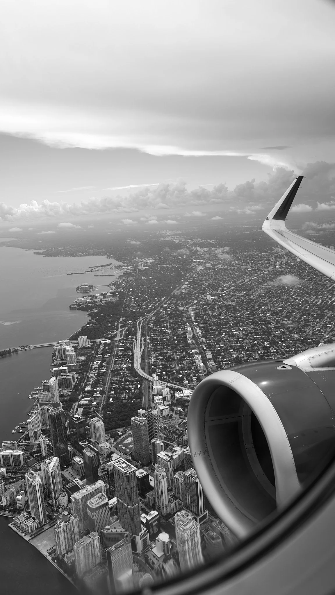 Black and white photo of a city and water taken from an airplane window, with part of the airplane wing visible.