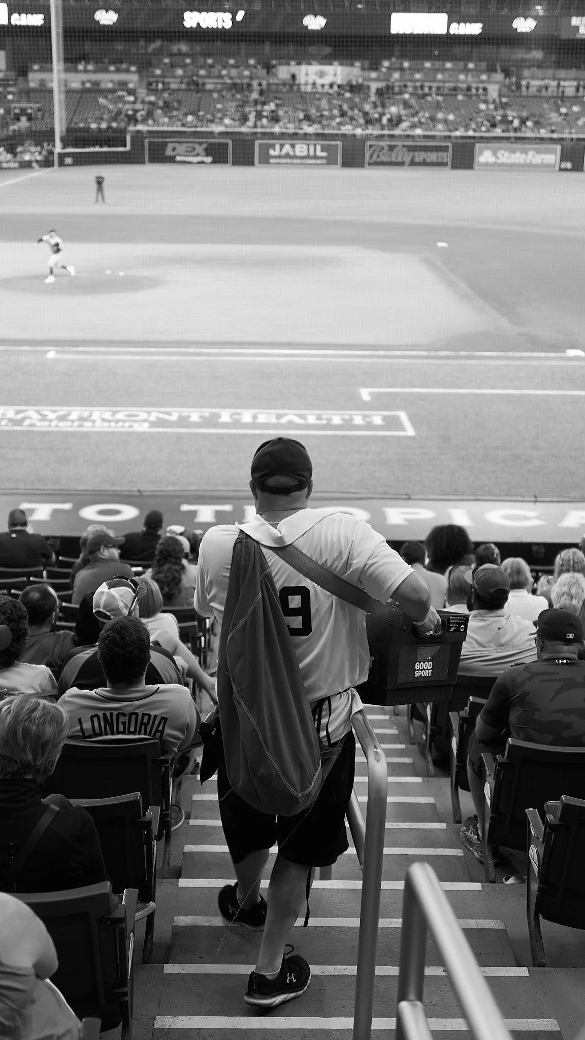 A baseball stadium with a man wearing a baseball jersey and backpack walking down the stairs toward the field, with spectators seated and a player on the field preparing to throw.
