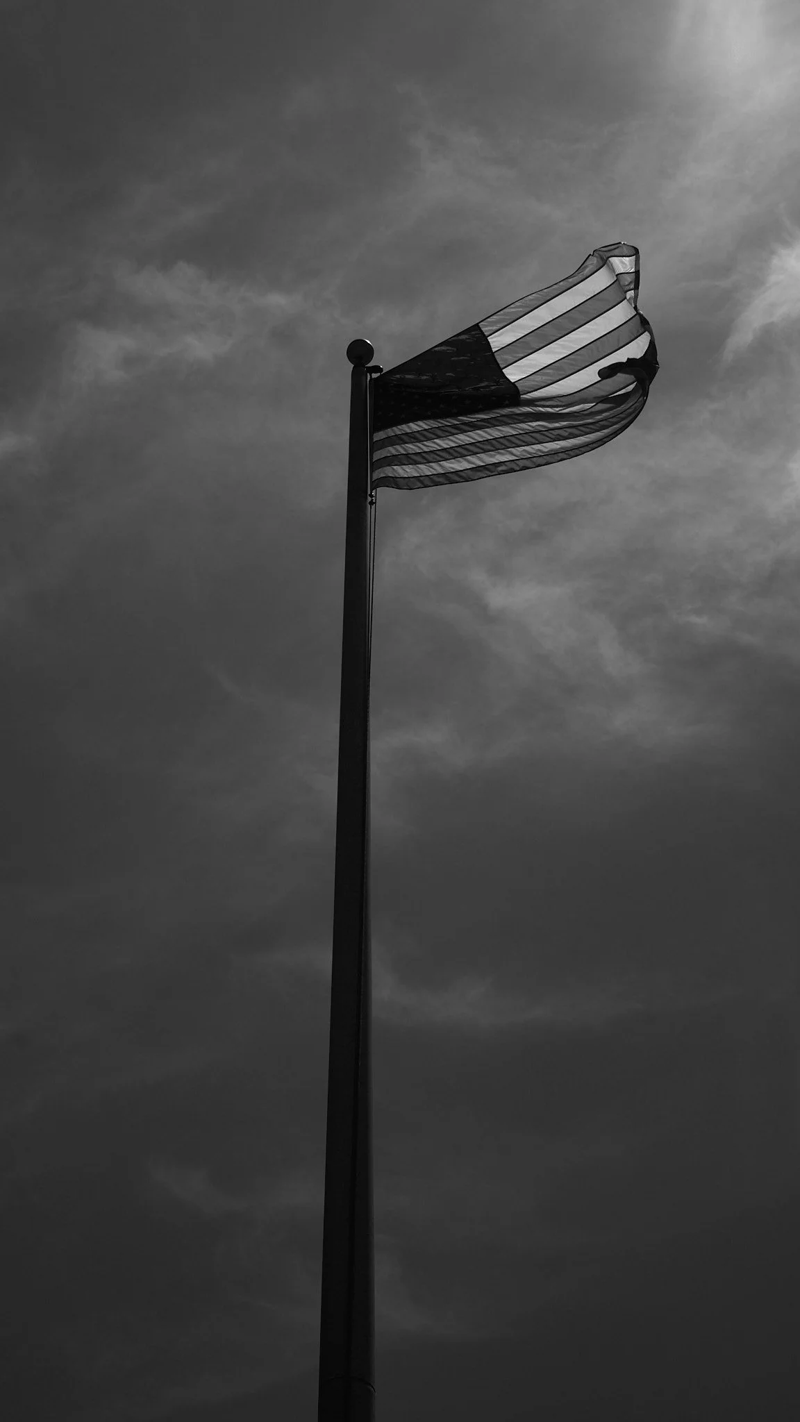 Black and white photo of an American flag waving on a flagpole against a cloudy sky.