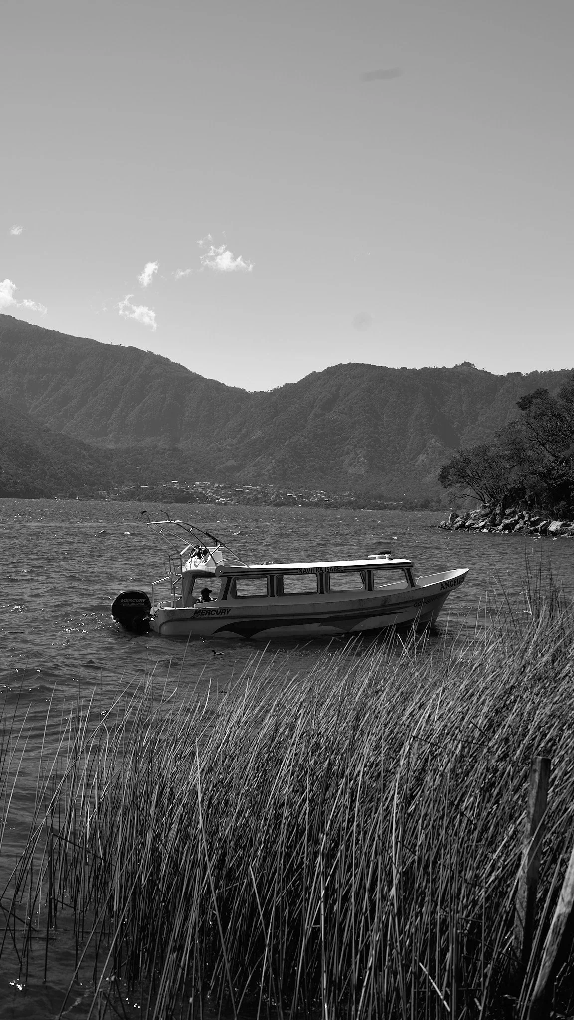 A black and white photo of a motorboat anchored near the grassy shoreline of a lake, with mountains and a clear sky in the background.