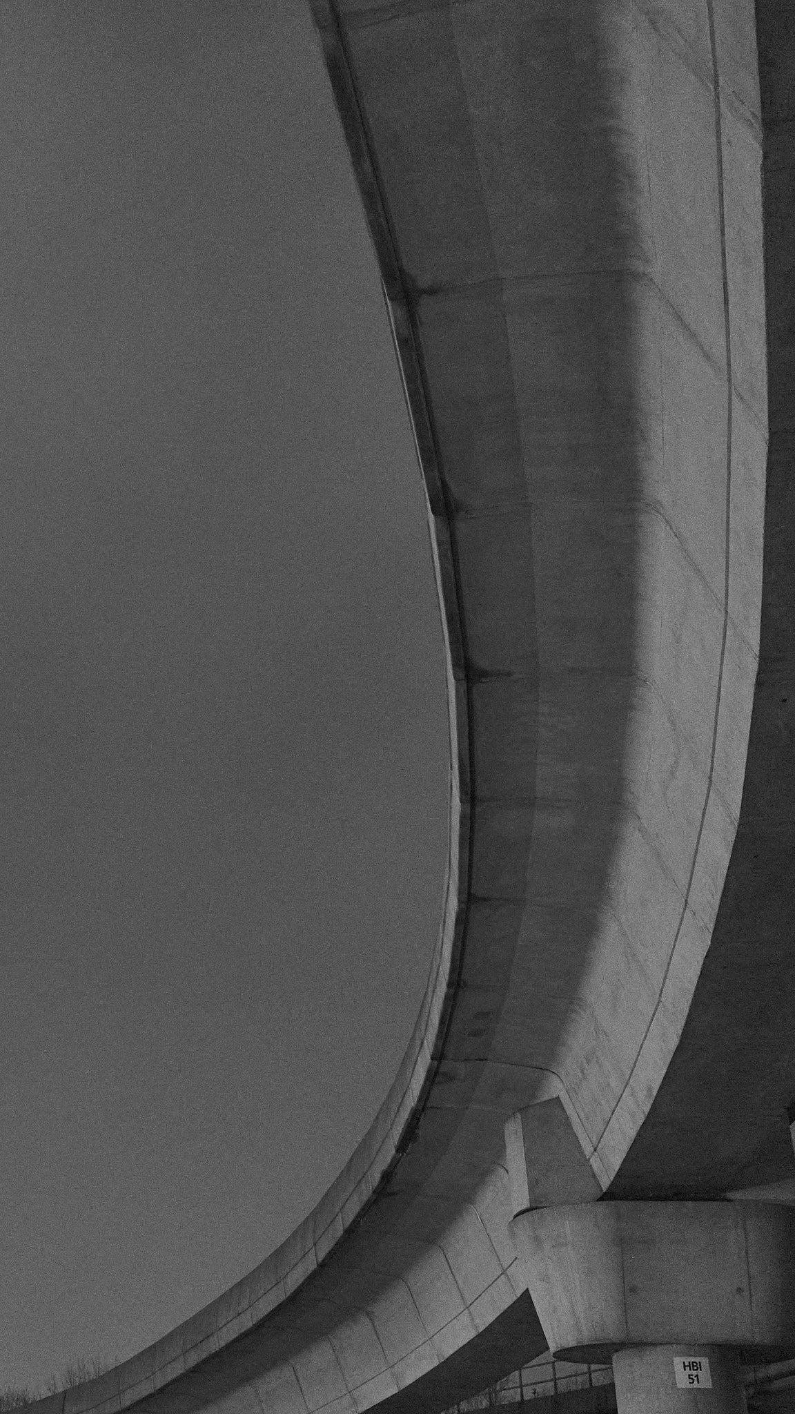 Black and white photo of a concrete bridge or overpass with a curved design, showing the underside and a support pillar.