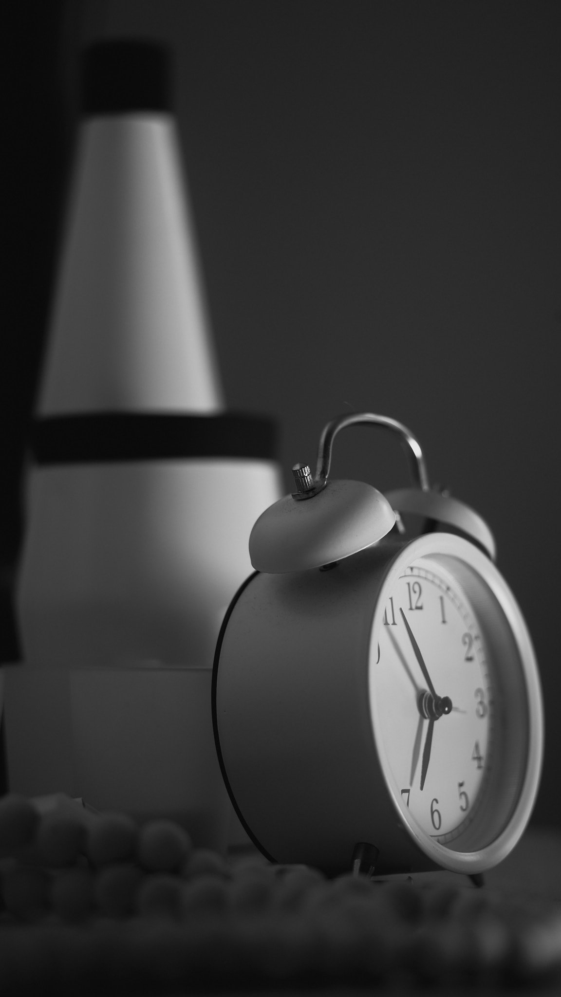 A black and white photo of an alarm clock showing 5:53, with a lamp in the background and small round objects in the foreground.